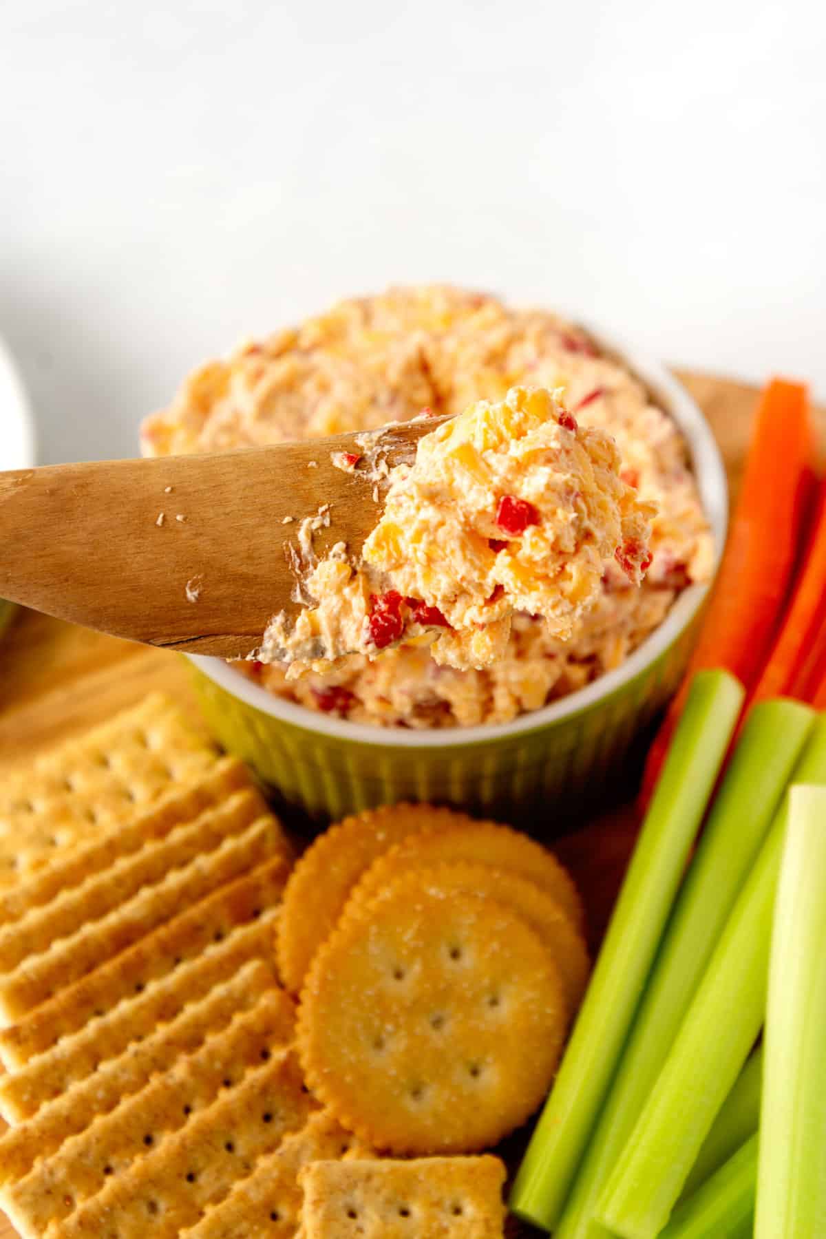 wooden spoonful of pimento cheese dip with a variety of crackers and cut vegetebles in the background sitting on a wooden board
