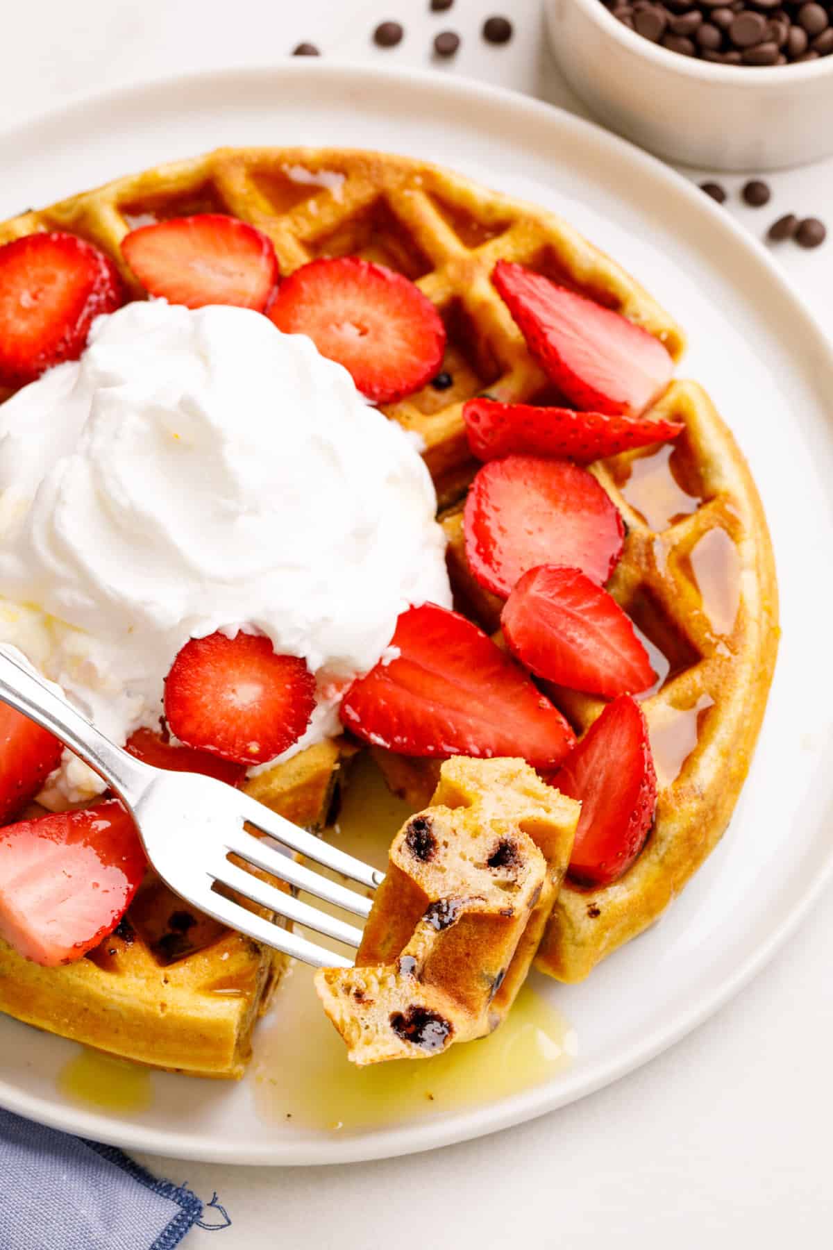 close up image of a bite of chocolate chip waffles topped with syrup, fresh strawberries, and whipped cream served on a white round plate