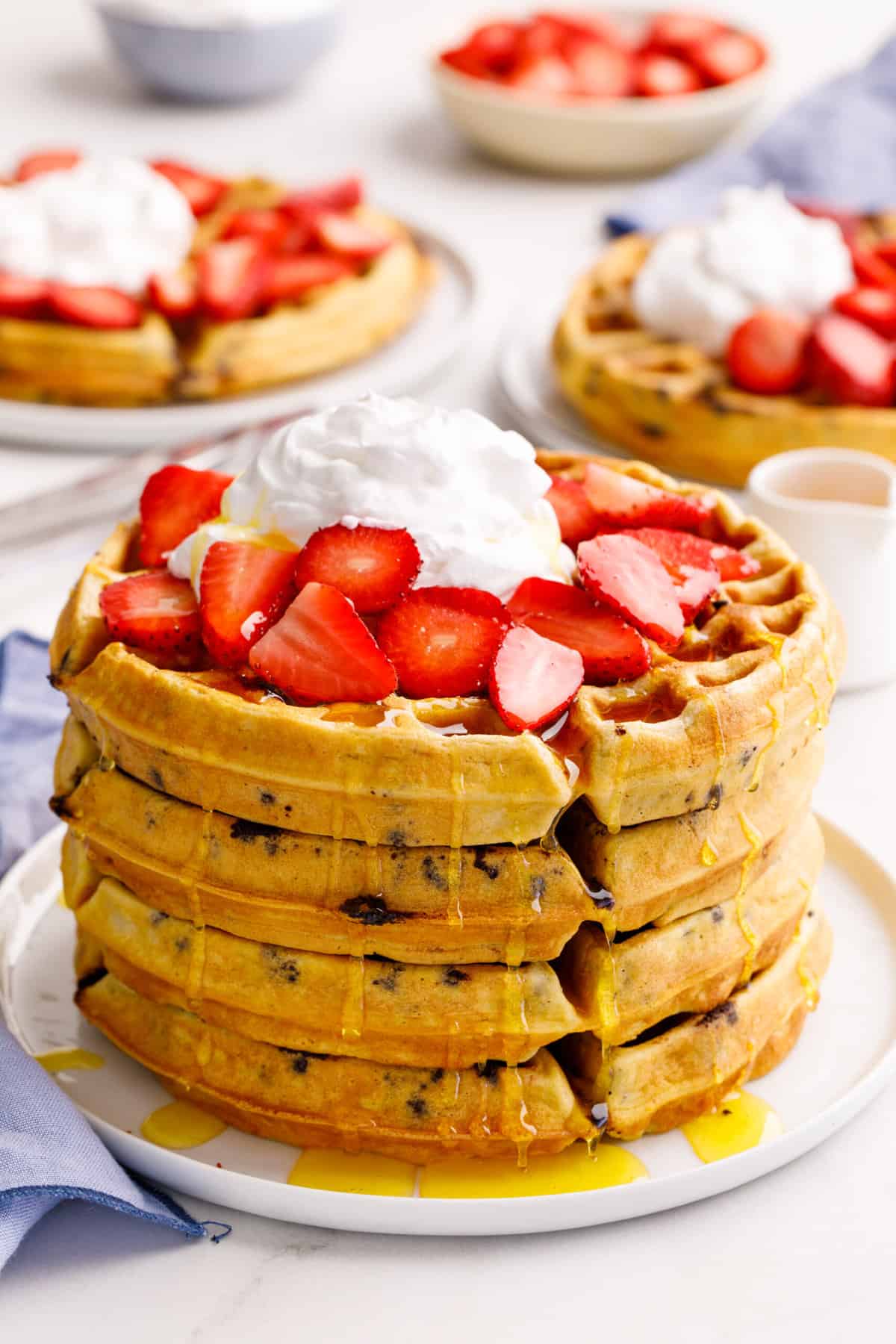stack of four chocolate chip waffles topped with syrup, fresh strawberries, and whipped cream served on a white round plate