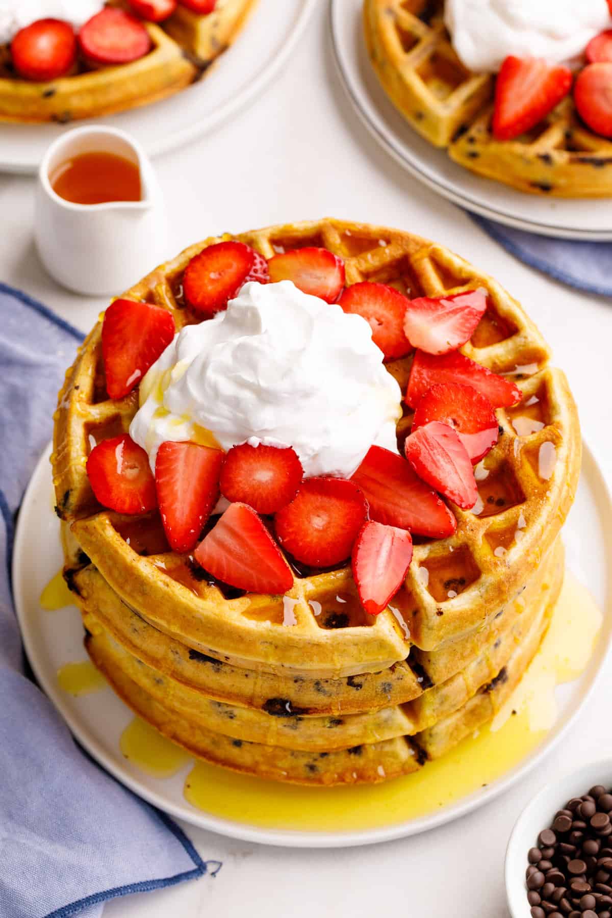 top down image of a stack of four chocolate chip waffles topped with syrup, fresh strawberries, and whipped cream served on a white round plate