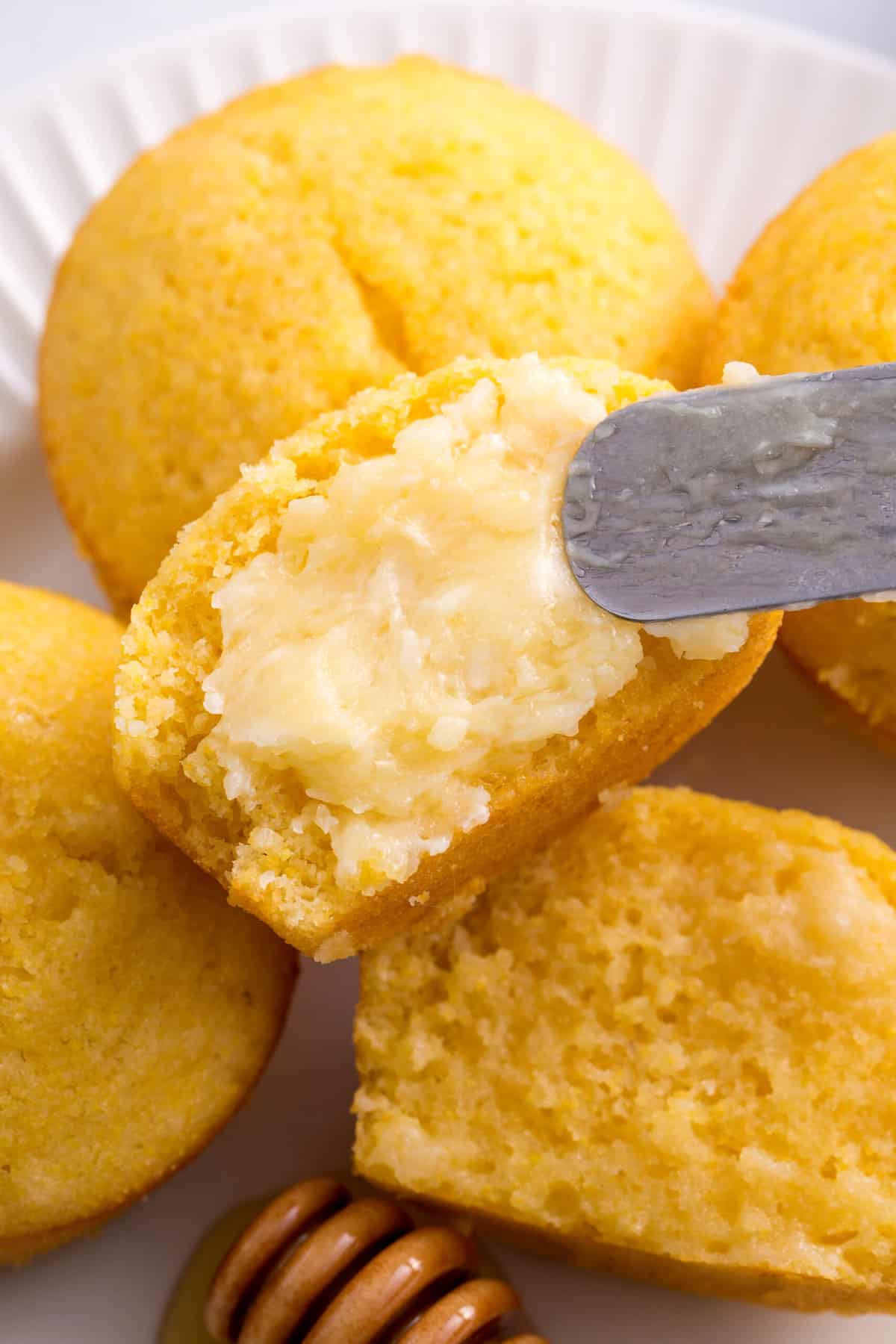 close up image of corn muffin halves spread with homemade honey butter with a metal spatula