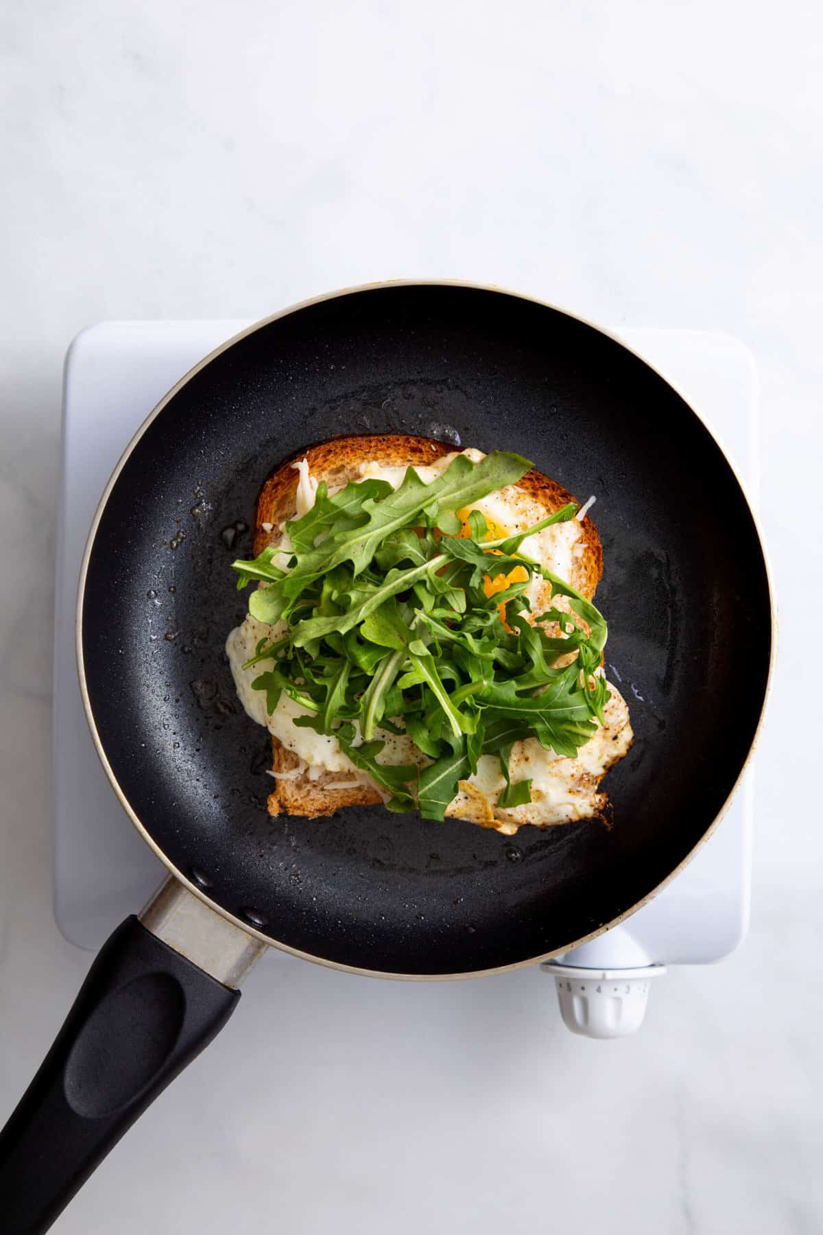 butter slice of white bread on a skillet topped with shredded cheese, a fried egg and fresh arugula