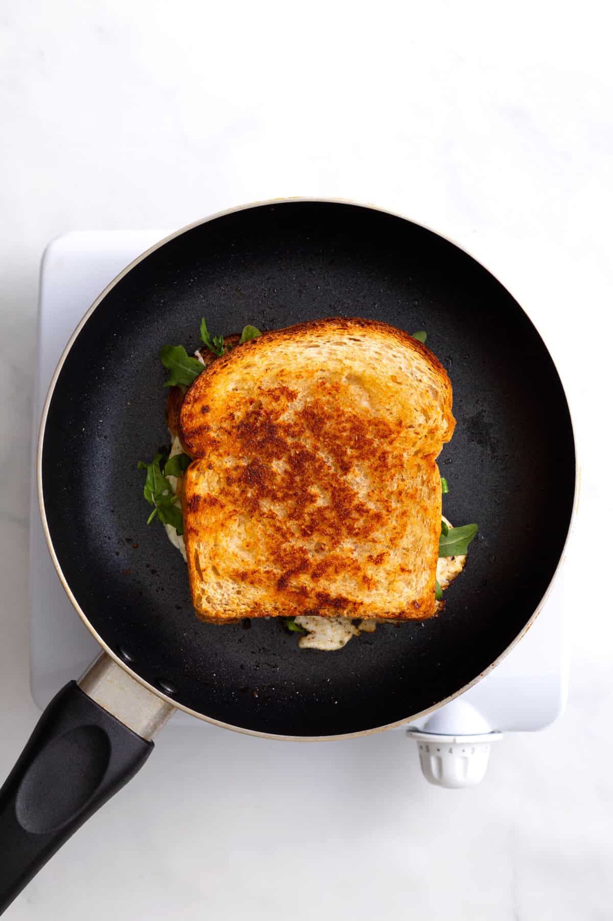 butter slice of white bread on a skillet topped with shredded cheese, a fried egg and fresh arugula and another slice of buttered toast to close the sandwich