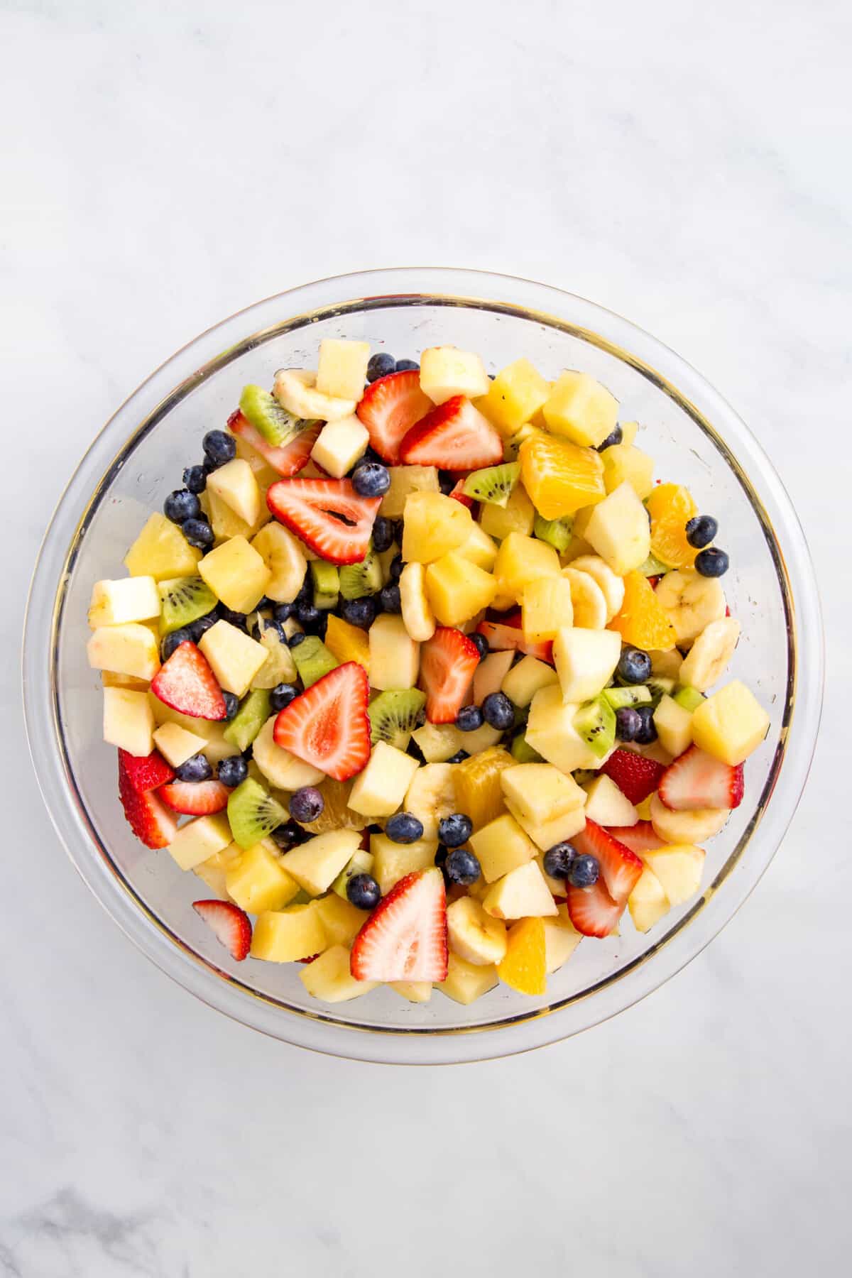 cut up strawberries, bananas, kiwi, oranges, pineapple, and fresh blueberries in a large glass bowl.