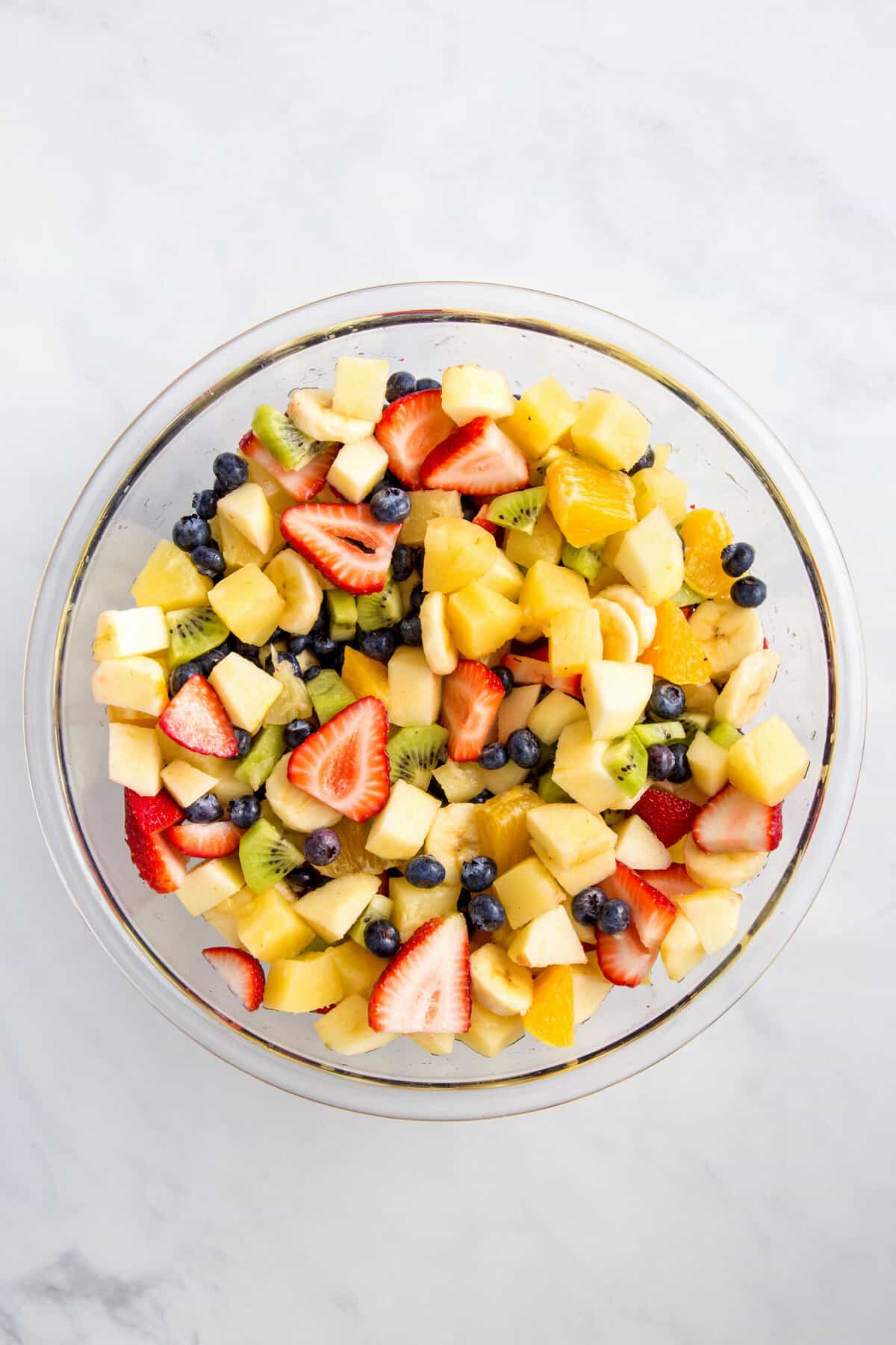 cut up strawberries, bananas, kiwi, oranges, pineapple, and fresh blueberries in a large glass bowl