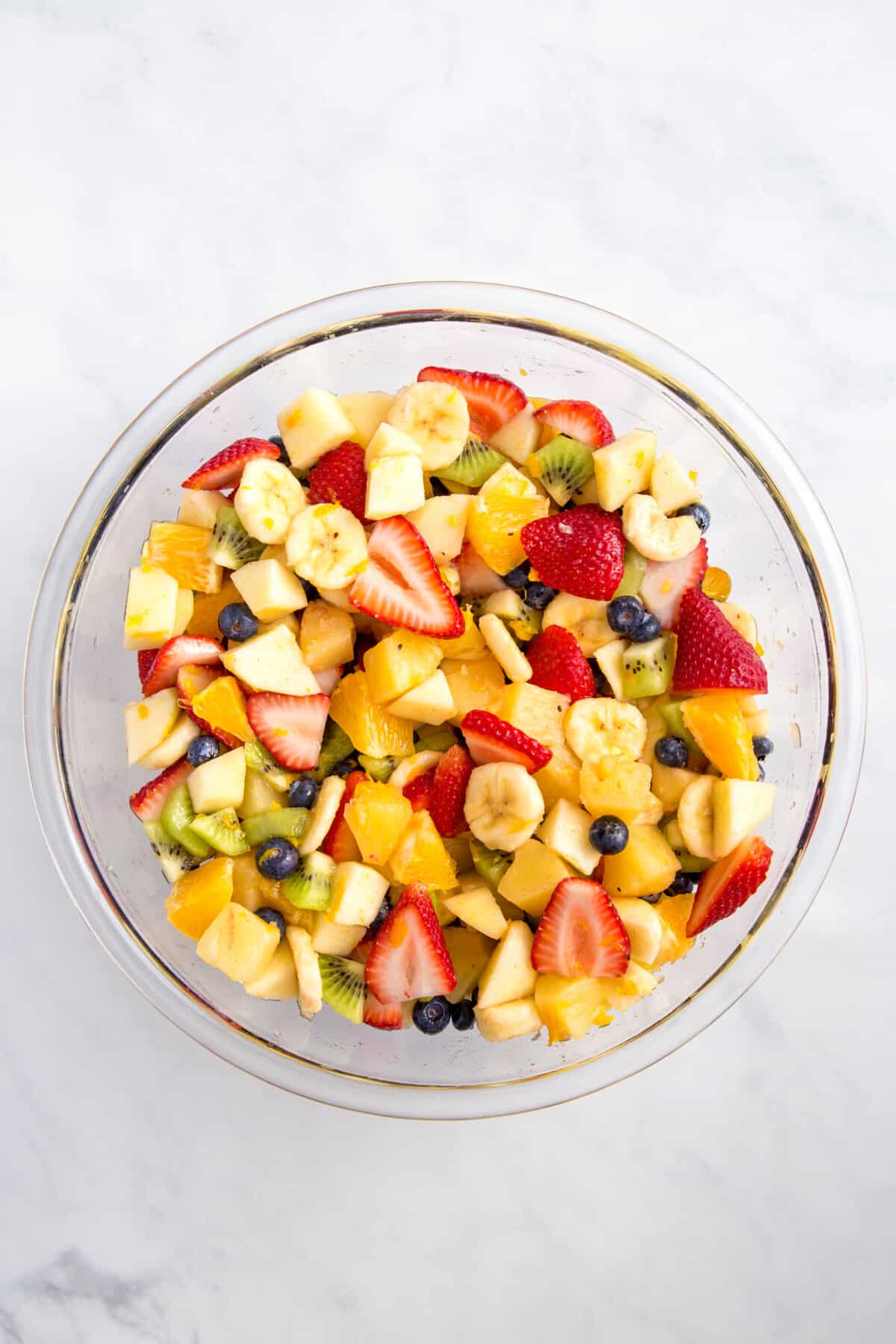cut up strawberries, bananas, kiwi, oranges, pineapple, and fresh blueberries tossed in dressing served in a large glass bowl.