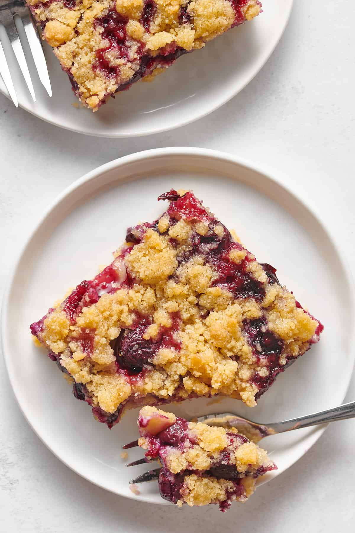 top down image of a square serving of blueberry crumble bar with a forkful of the dessert sitting on a white round plate