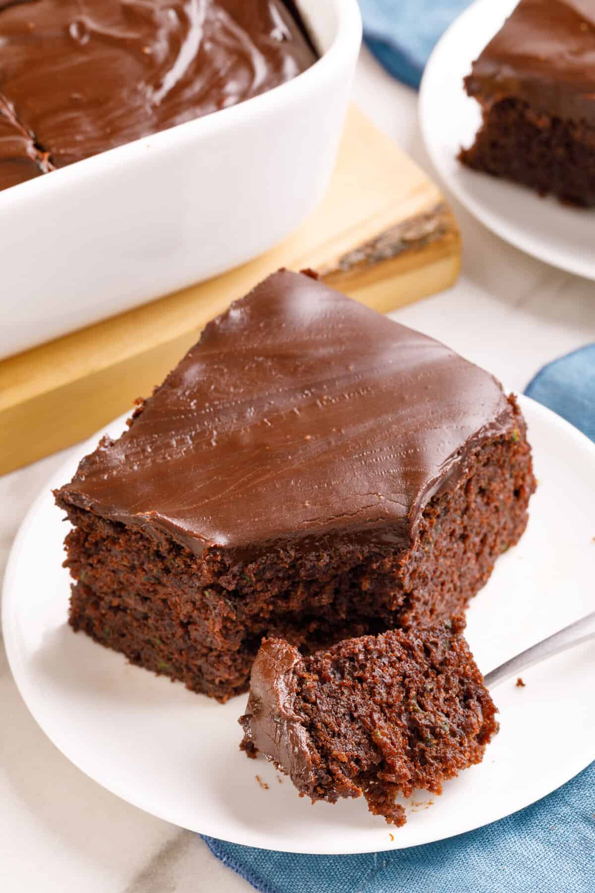 square slice of chocolate zucchini cake served on a white round plate with a forkful of cake