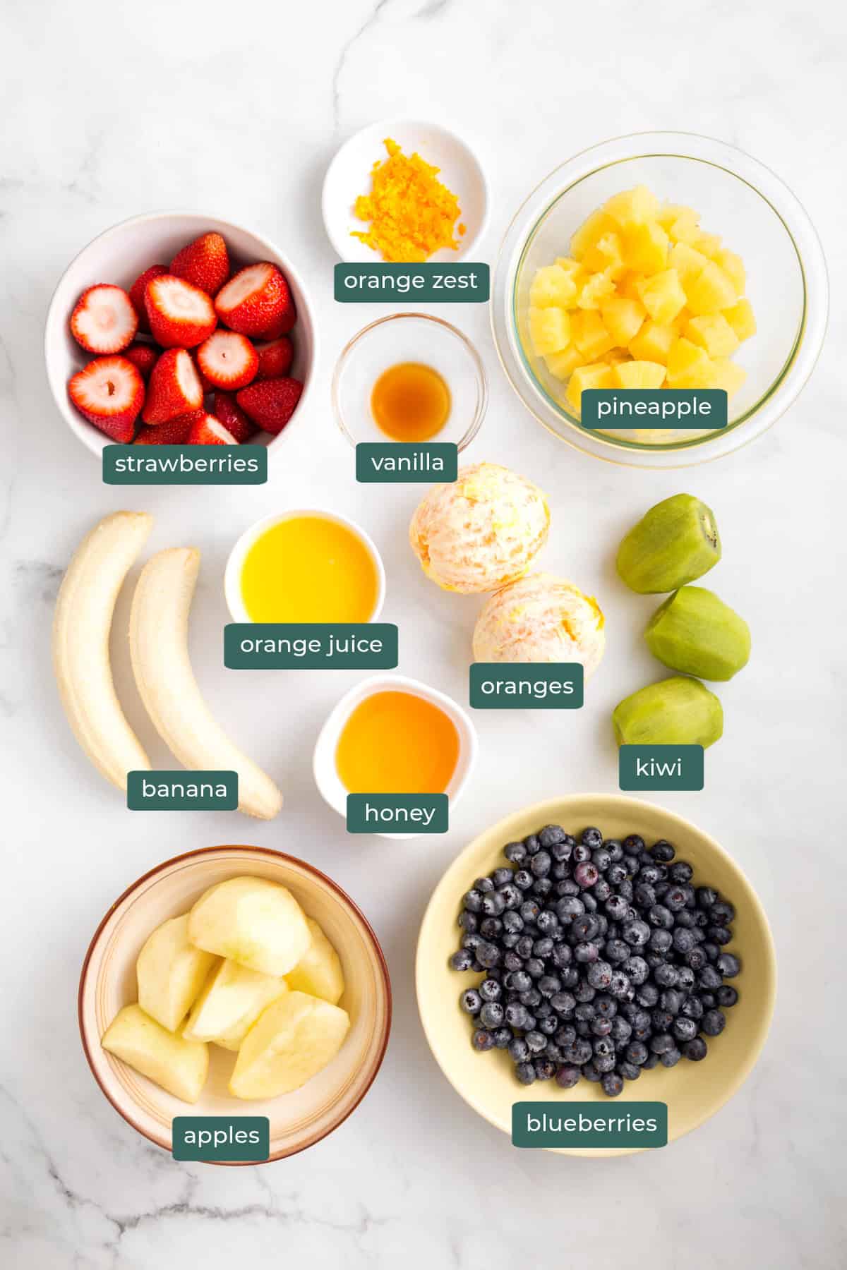 All of the ingredients for fruit salad in bowls on a white countertop.
