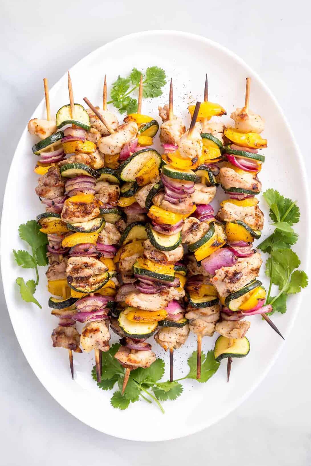 grilled chicken and vegetable kabobs sitting on a white oval plate garnished with cilantro