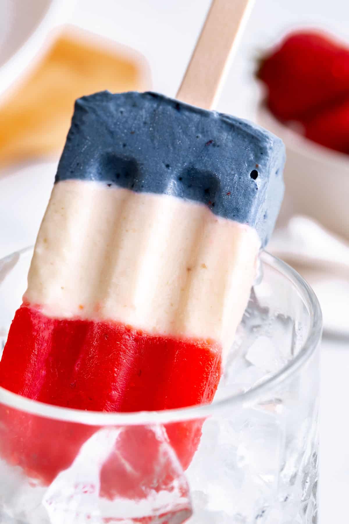 a red, white and blue popsicle facing down in a clear glass with ice cubes