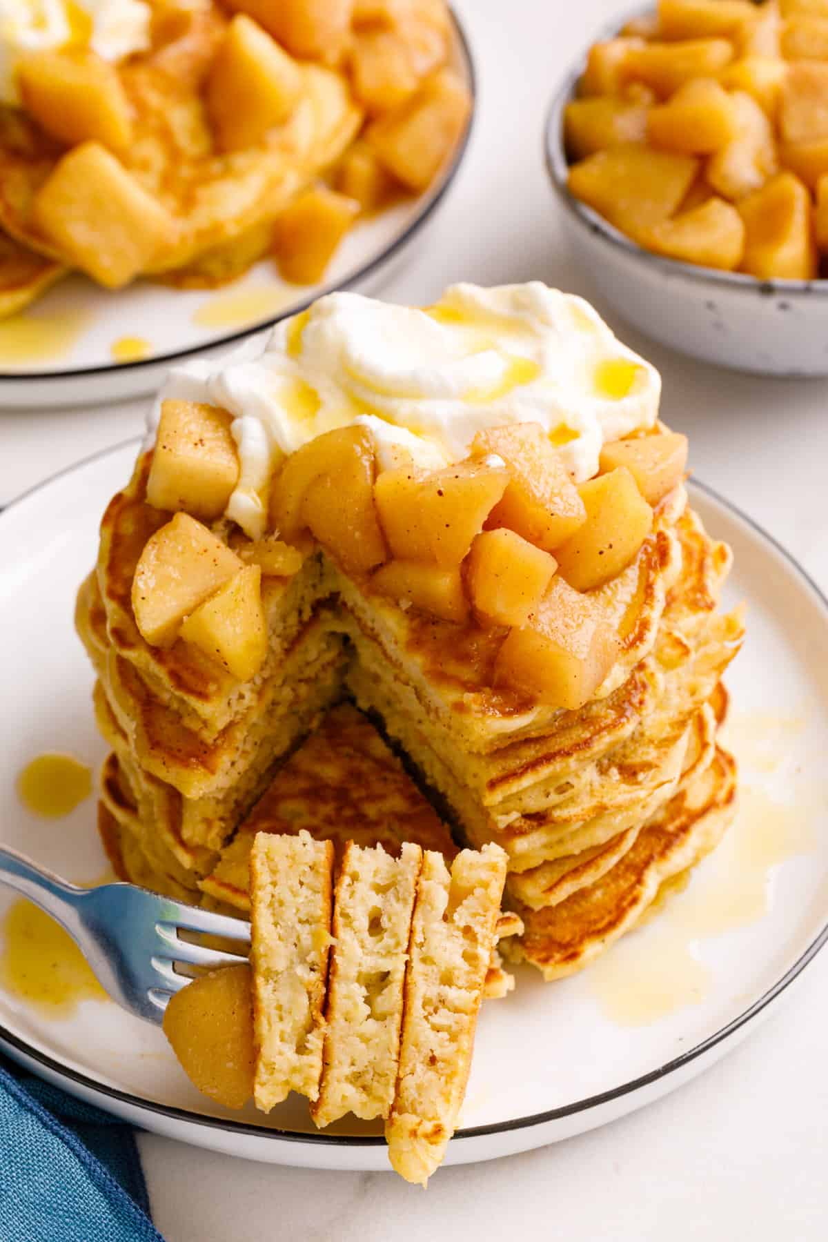 stack of four apple pancakes topped with fresh cooked granny smith apples, whipped cream and syrup with a slice taken out to show the cross section.