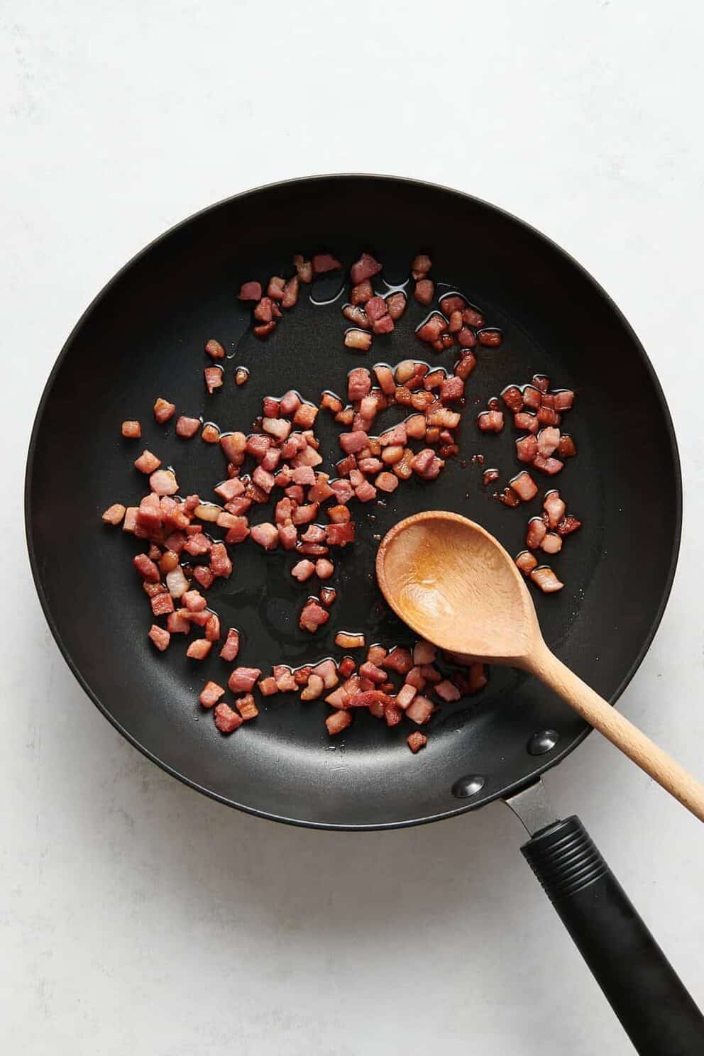 diced bacon cooking in a pan with a wooden spoon