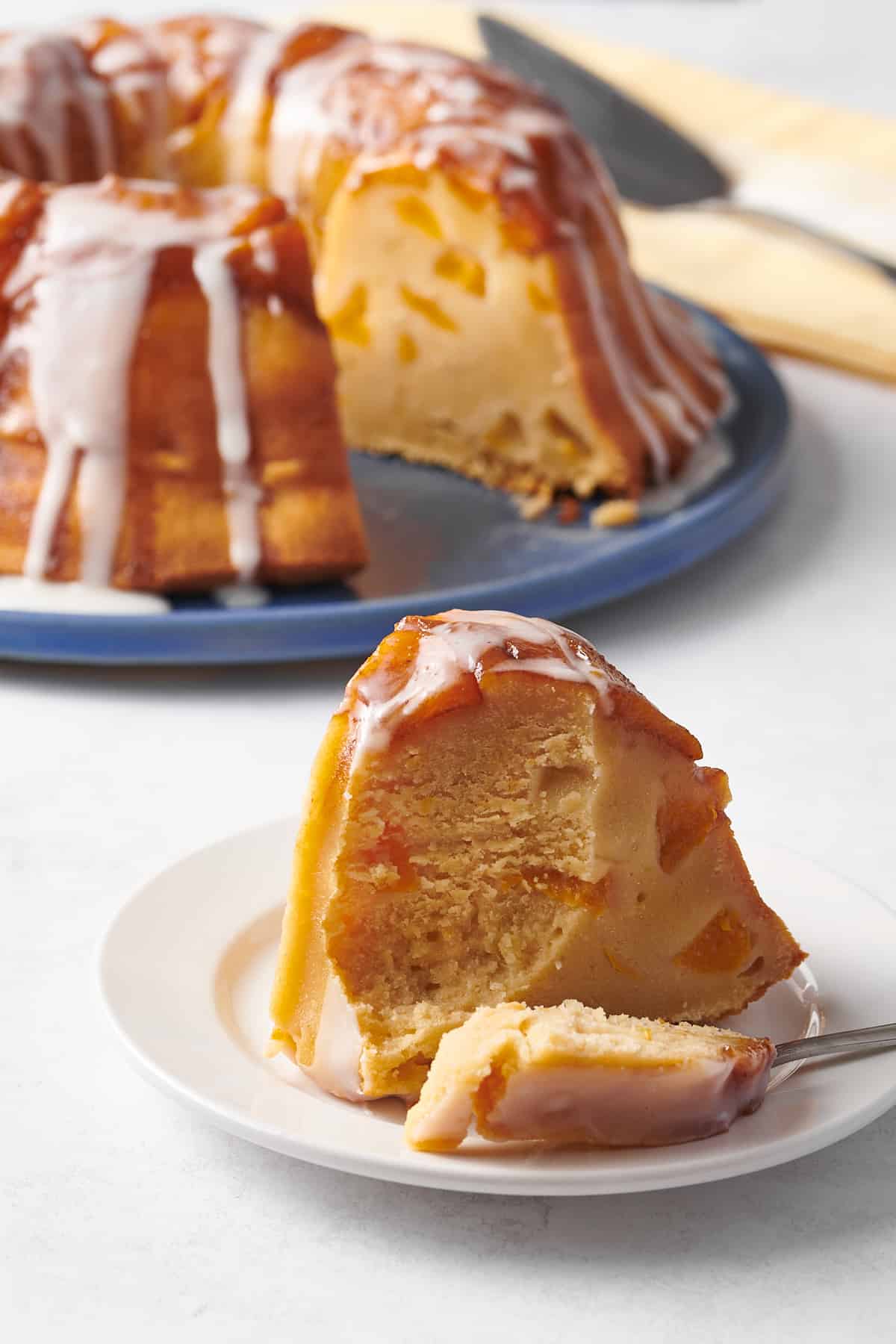 slice of peach cobbler pound cake sitting on a white round plate with the rest of the bundt pound cake in the background