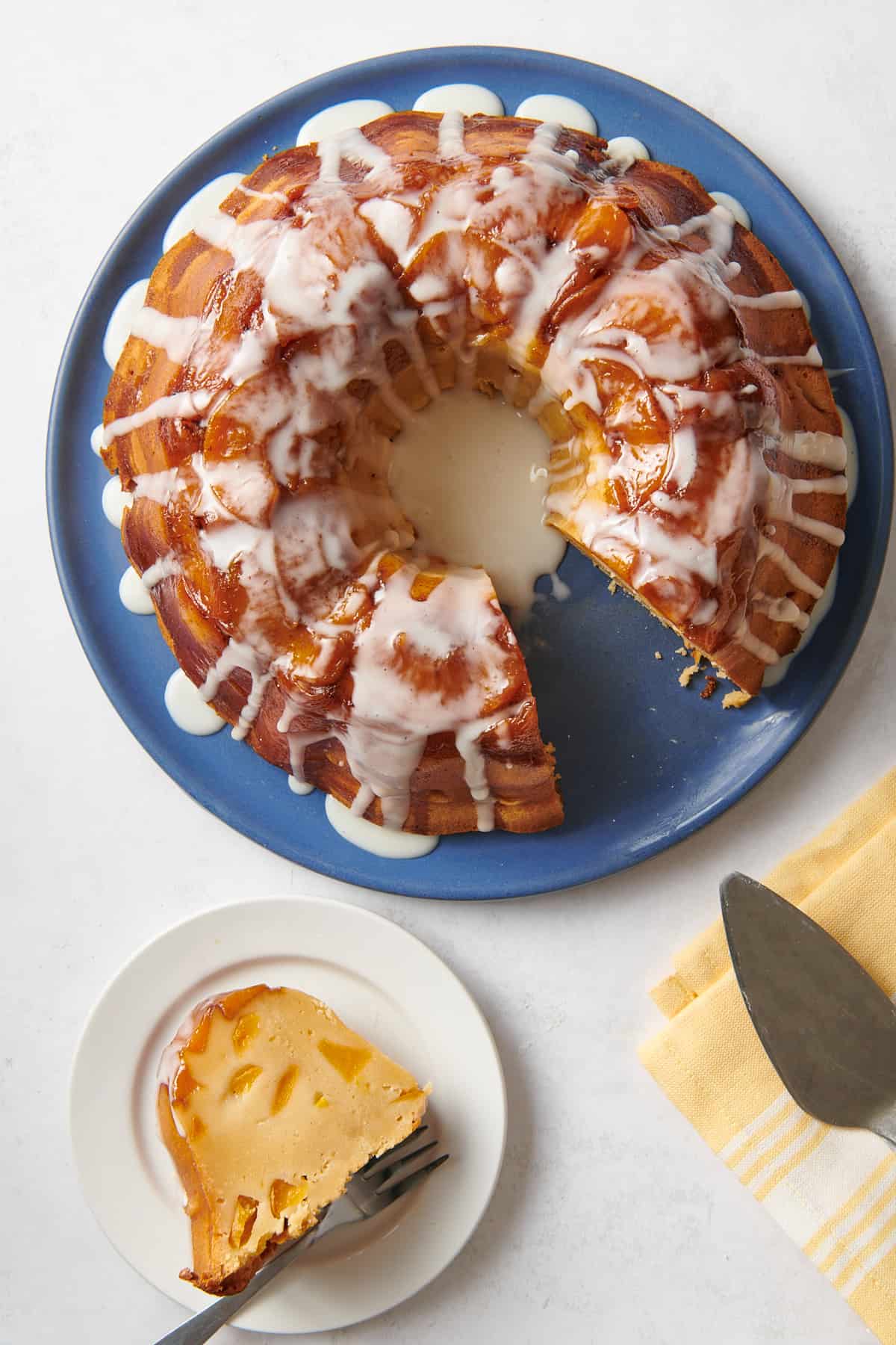 top down image of a slice of peach cobbler pound cake sitting on a white round plate with a dark silver fork and the rest of the bundt cake served on a large dark blue plate