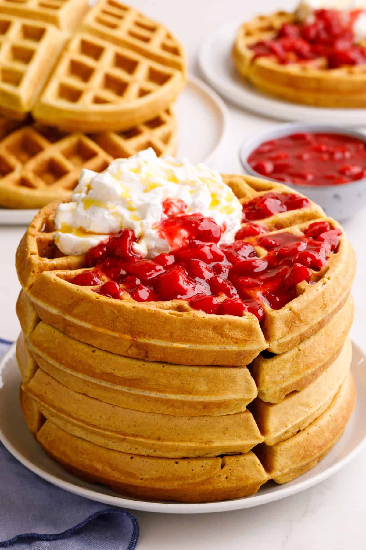 stack of four sourdough waffles topped with strawberry compote and whipped cream served on a white round plate