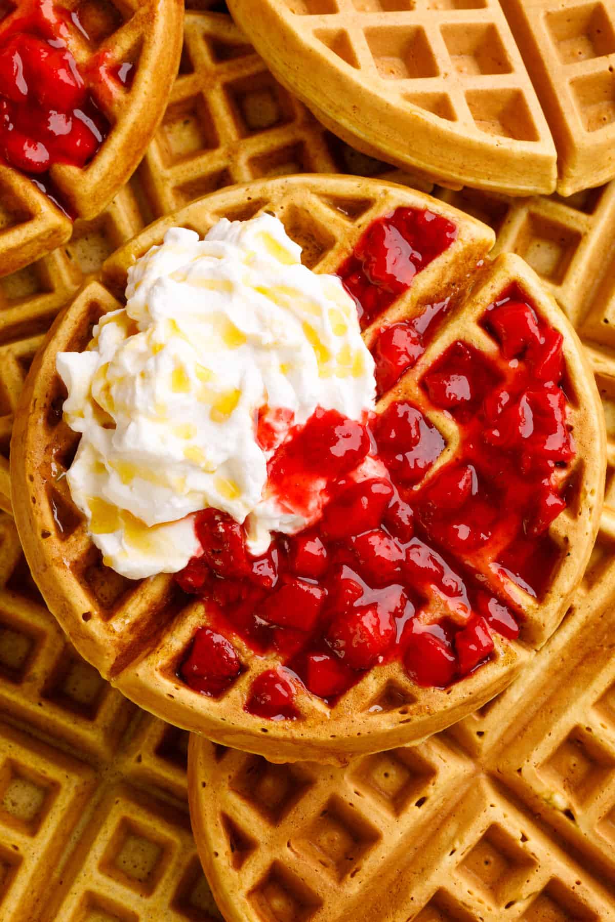 top down image of a sourdough waffle topped with strawberry compote and whipped cream sitting on a pile of sourdough waffles