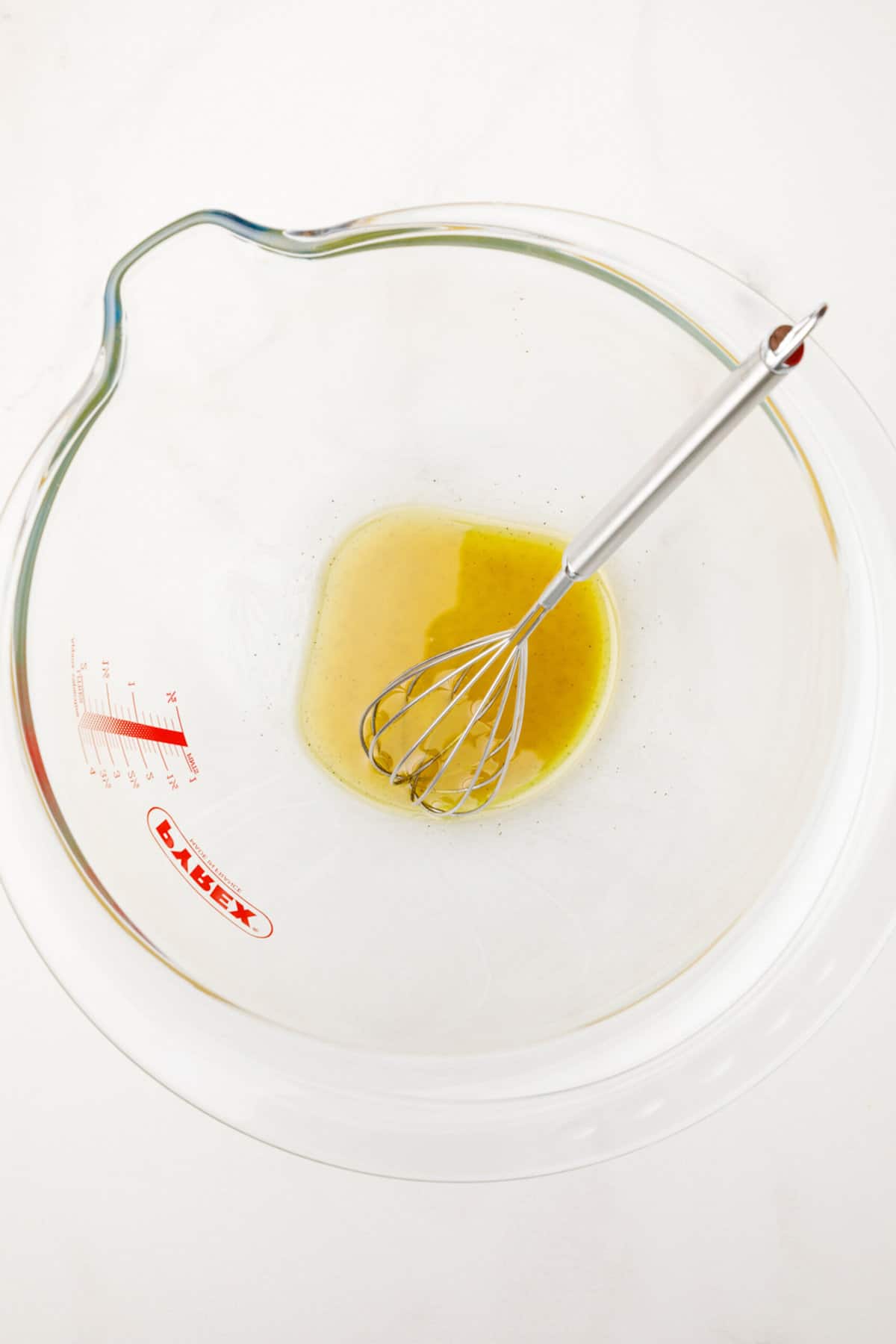 olive oil, vinegar, salt and pepper dressing sitting in a large clear mixing bowl with a metal whisk