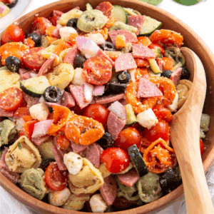 A bowl of tortellini pasta salad with a wooden spoon on the side.