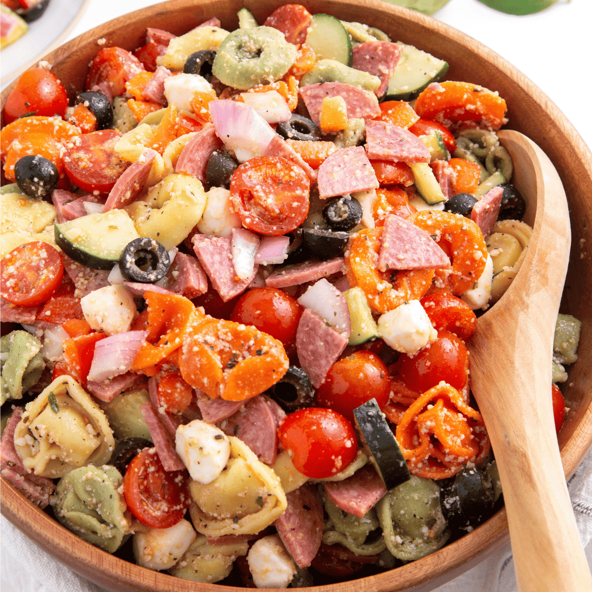 A bowl of tortellini pasta salad with a wooden spoon on the side.