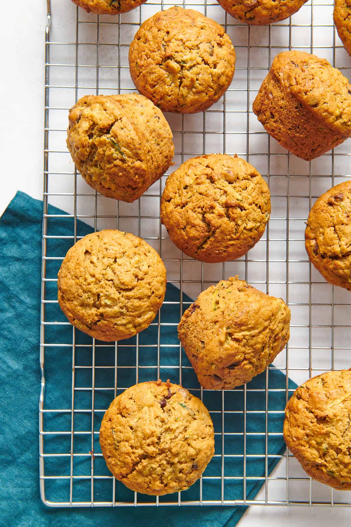 top down image of several zucchini muffins sitting on a wire cooling rack with a dark teal kitchen towel underneath the rack