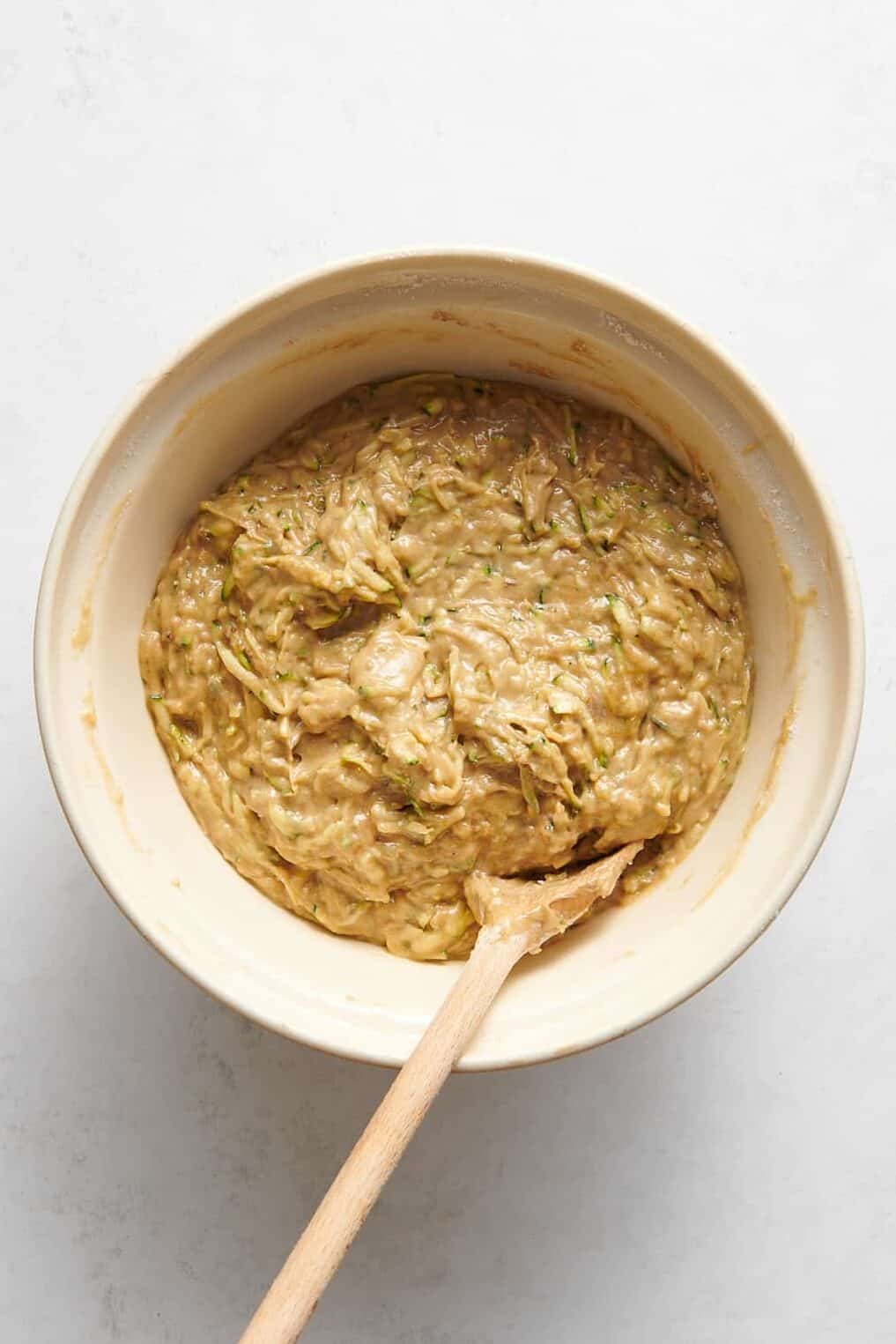 zucchini muffin batter sitting in a ceramic mixing bowl with a wooden spoon