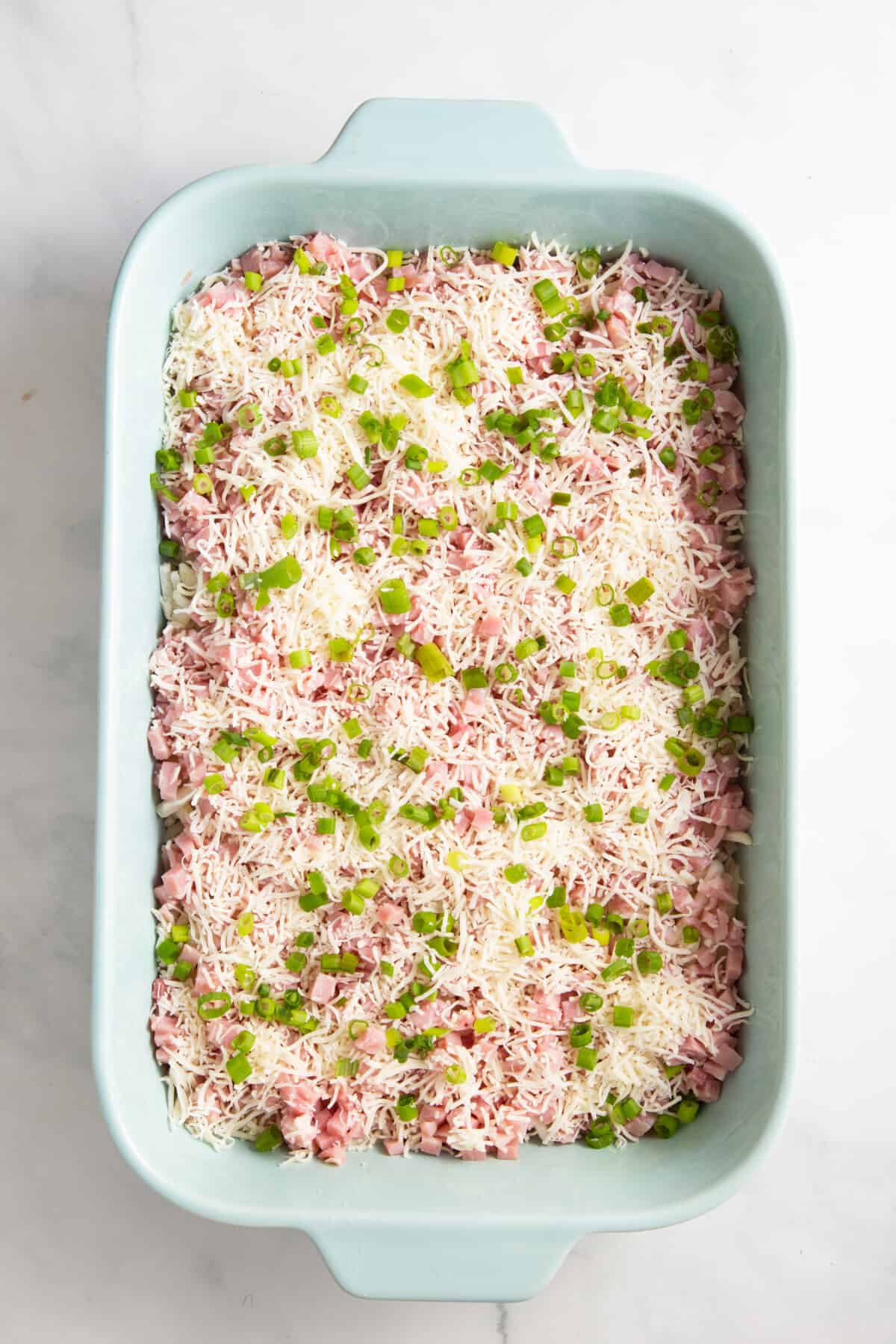 light blue 9x13 casserole dish with frozen hashbrowns, chopped ham, shredded cheese and chopped green onions