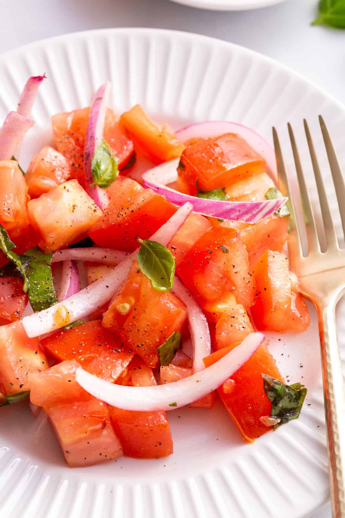 close up image of a serving of tomato salad sitting on a white round plate with a gold fork