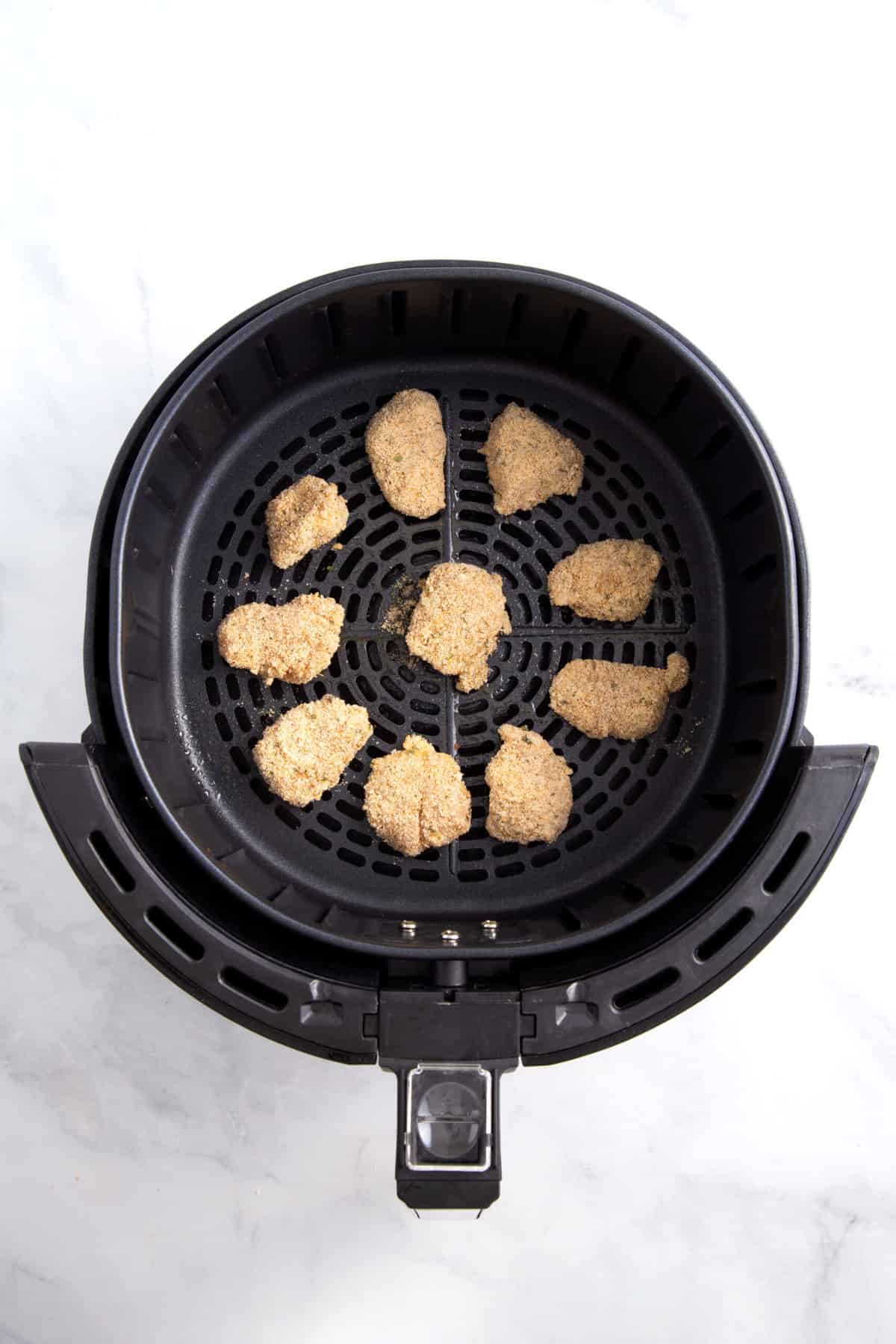 nugget sized chicken pieces coated in flour and dredged in an egg mixture, then coated with  a breadcrumb mixture sitting in an air fryer