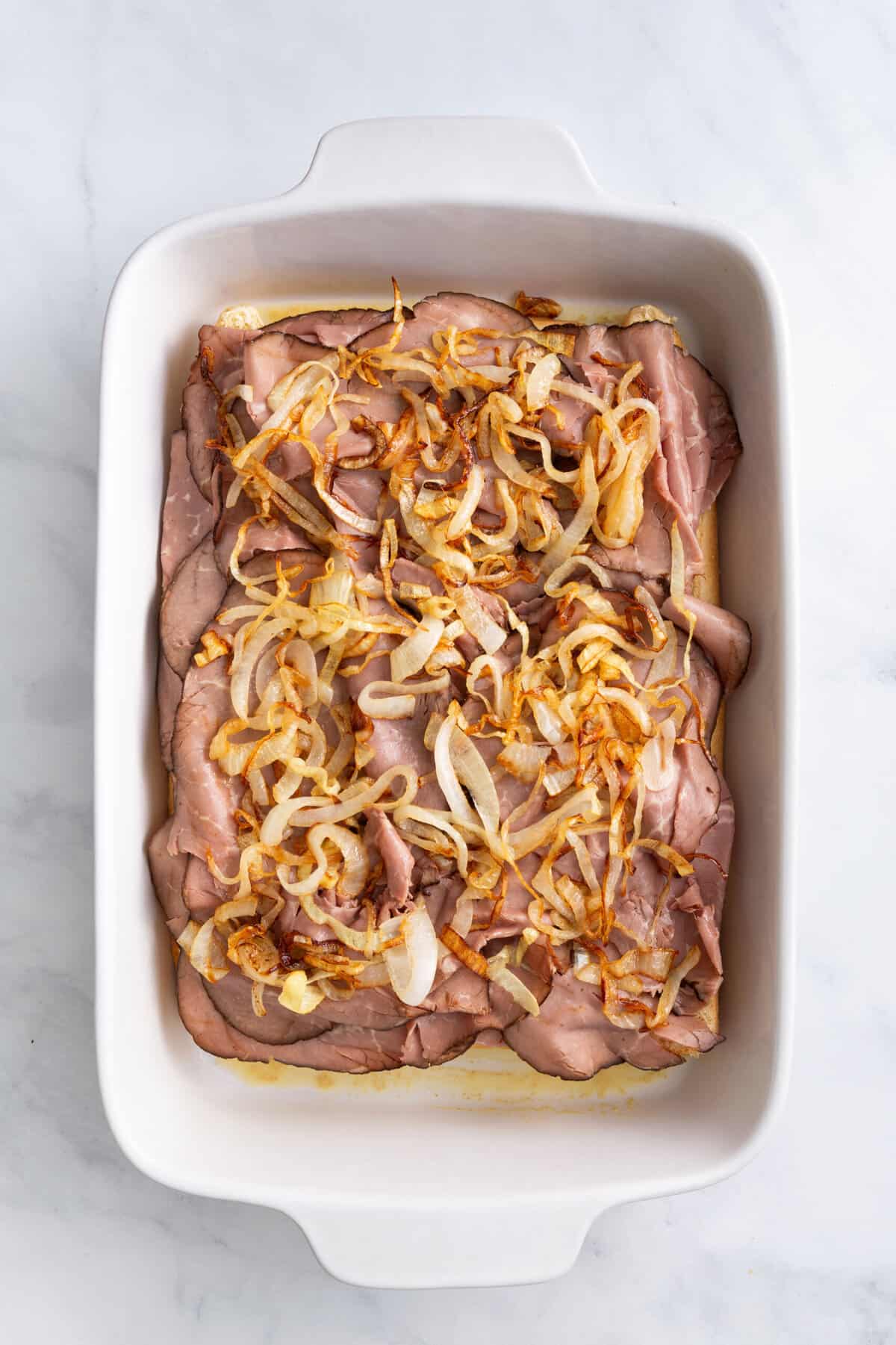 hawaiian roles arranged at the bottom of a 9x13 casserole dish with thinly sliced roast beef arranged on top of the bread evenly and cooked caramelized onions on top