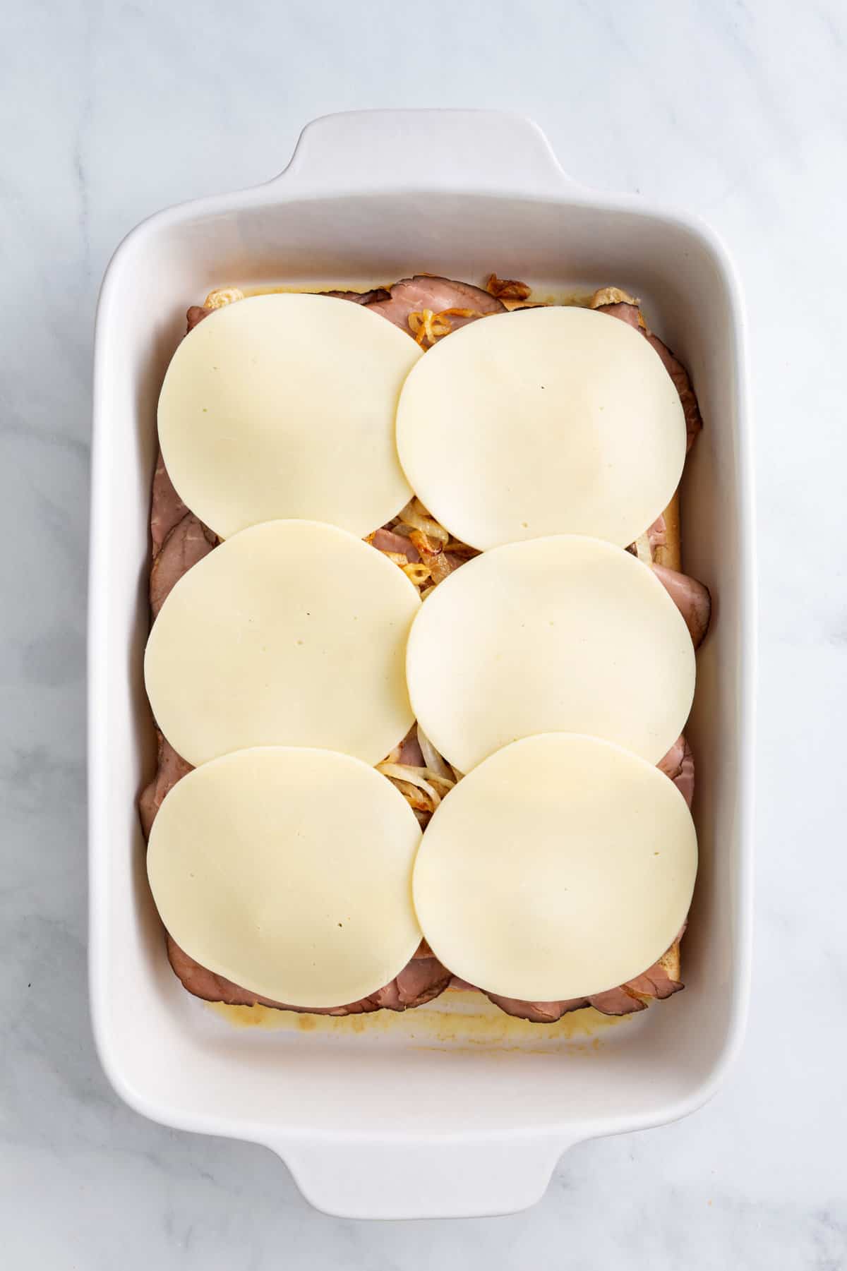 hawaiian roles arranged at the bottom of a 9x13 casserole dish with thinly sliced roast beef arranged on top of the bread evenly and cooked caramelized onions and topped with a layer of sliced provolone cheese