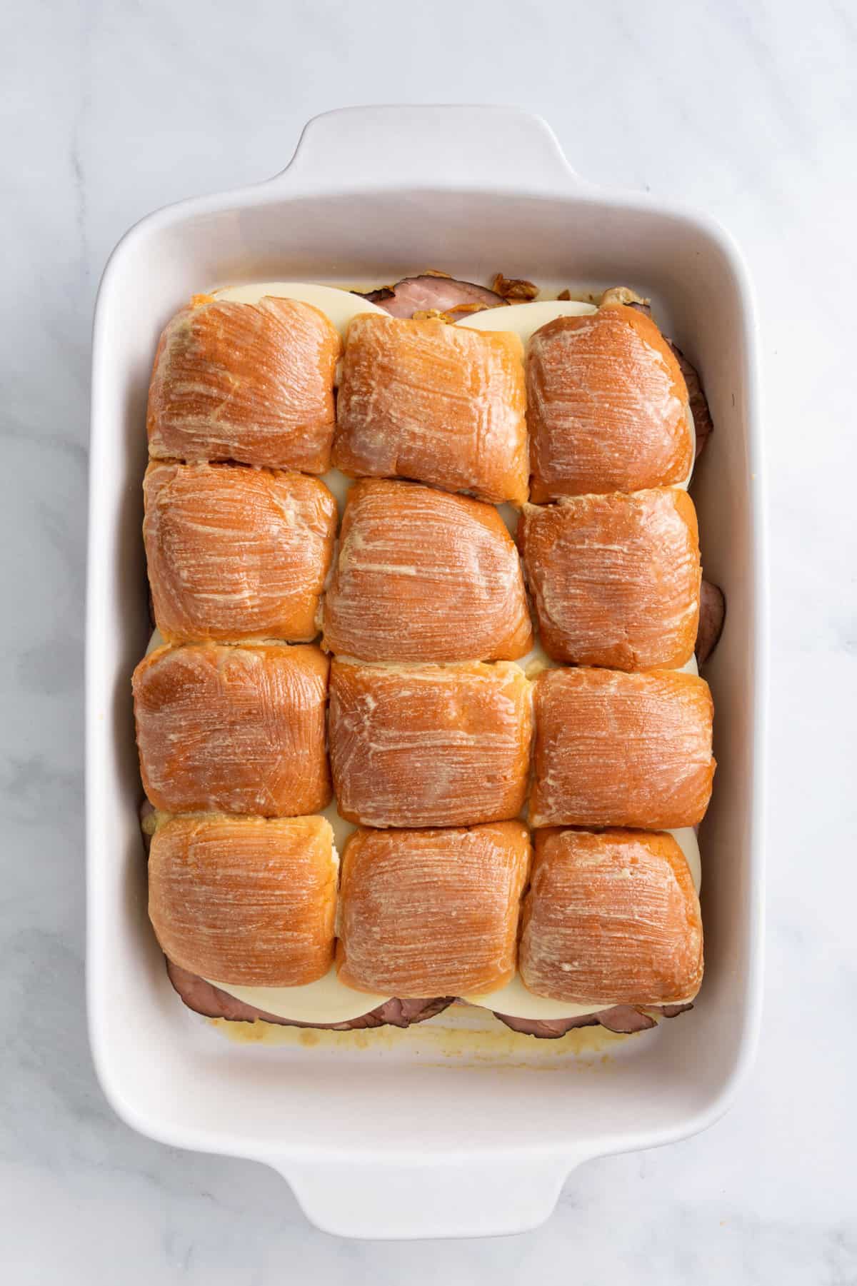 hawaiian roles arranged at the bottom of a 9x13 casserole dish with thinly sliced roast beef arranged on top of the bread evenly and cooked caramelized onions and a layer of sliced provolone cheese then topped with the hawaiian rolls bread and spread with softened butter