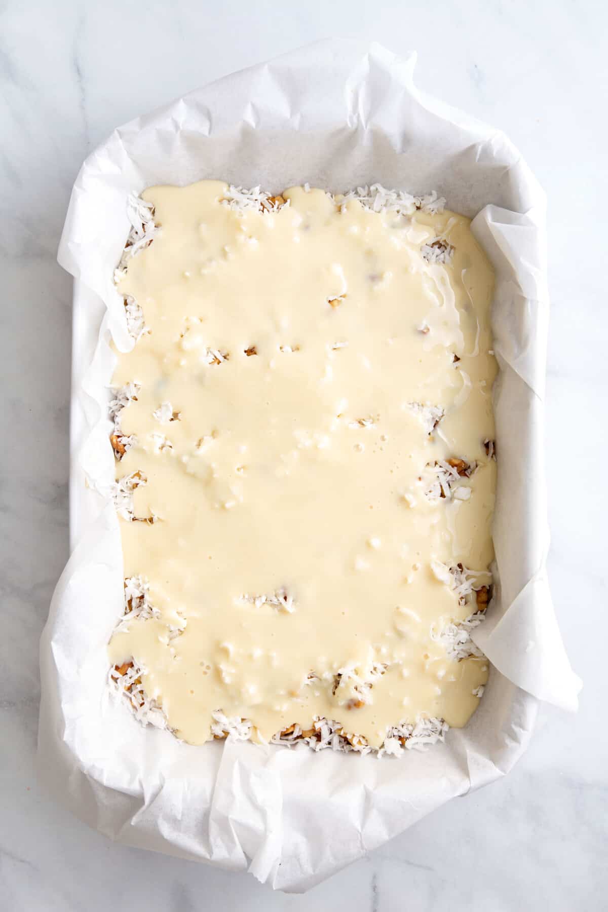 graham cracker crust pressed into a parchment paper lined 9x13 baking dish topped with a layer of milk and white chocolate chips, chopped pecans, and shredded coconut drizzled with sweetened condensed milk on top