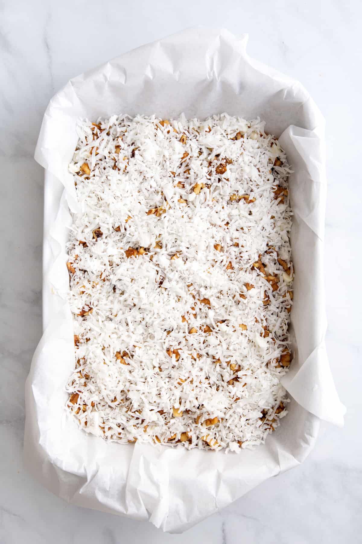 graham cracker crust pressed into a parchment paper lined 9x13 baking dish topped with a layer of milk and white chocolate chips, chopped pecans, and shredded coconut