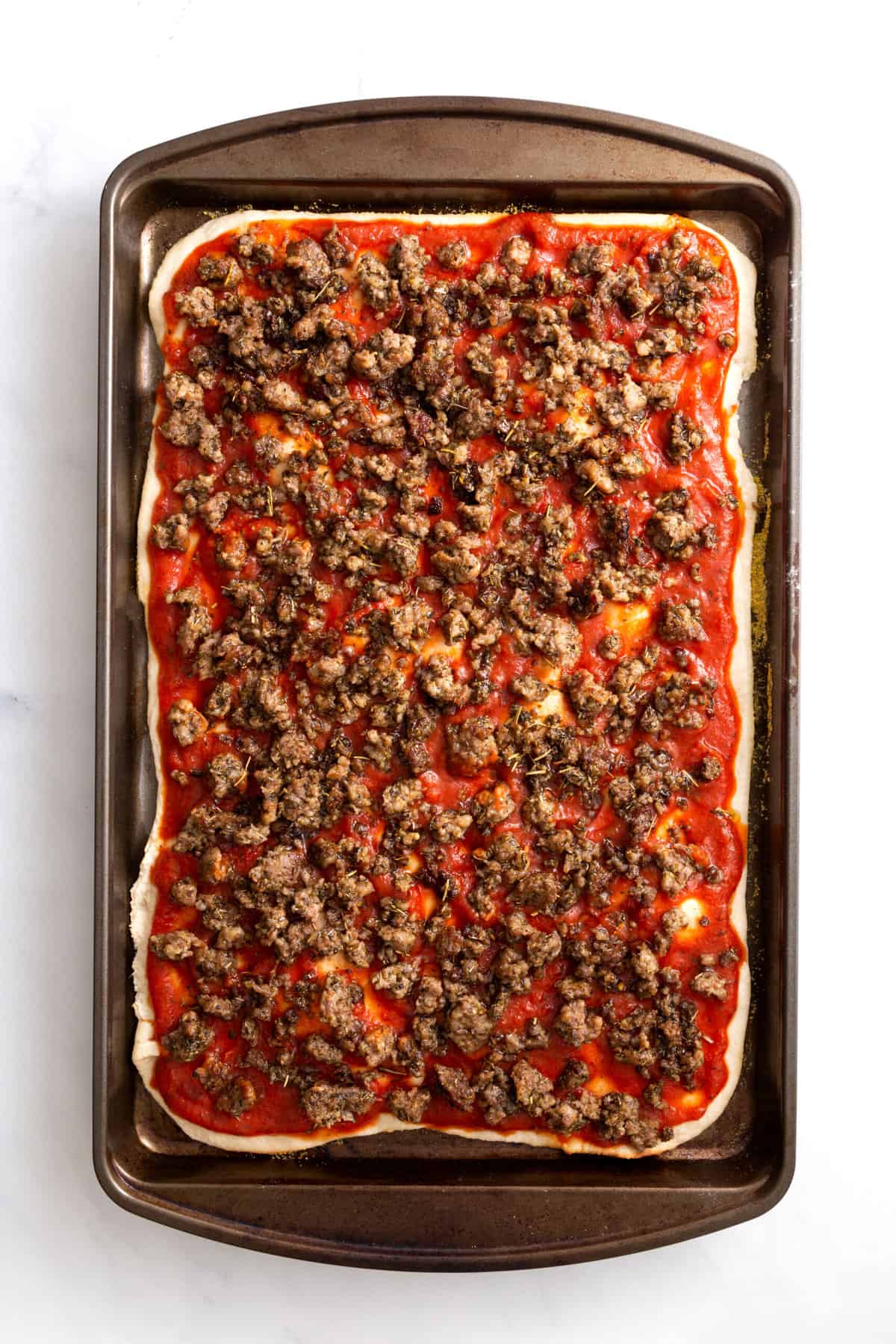pizza dough layered at the bottom of a sheet pan, with pizza sauce, cooked sausage