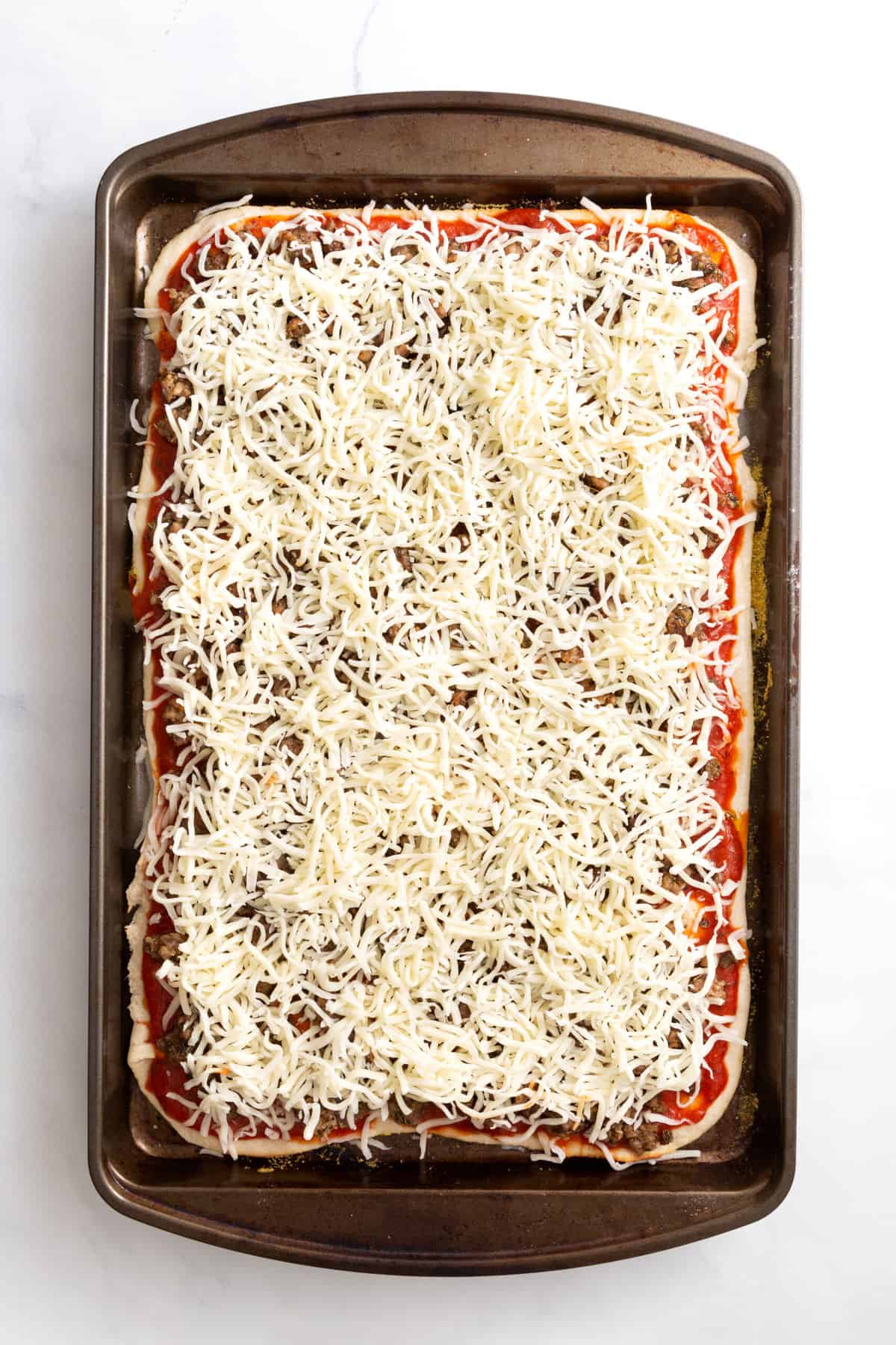 pizza dough layered at the bottom of a sheet pan, with pizza sauce, cooked sausage and shredded mozzarella