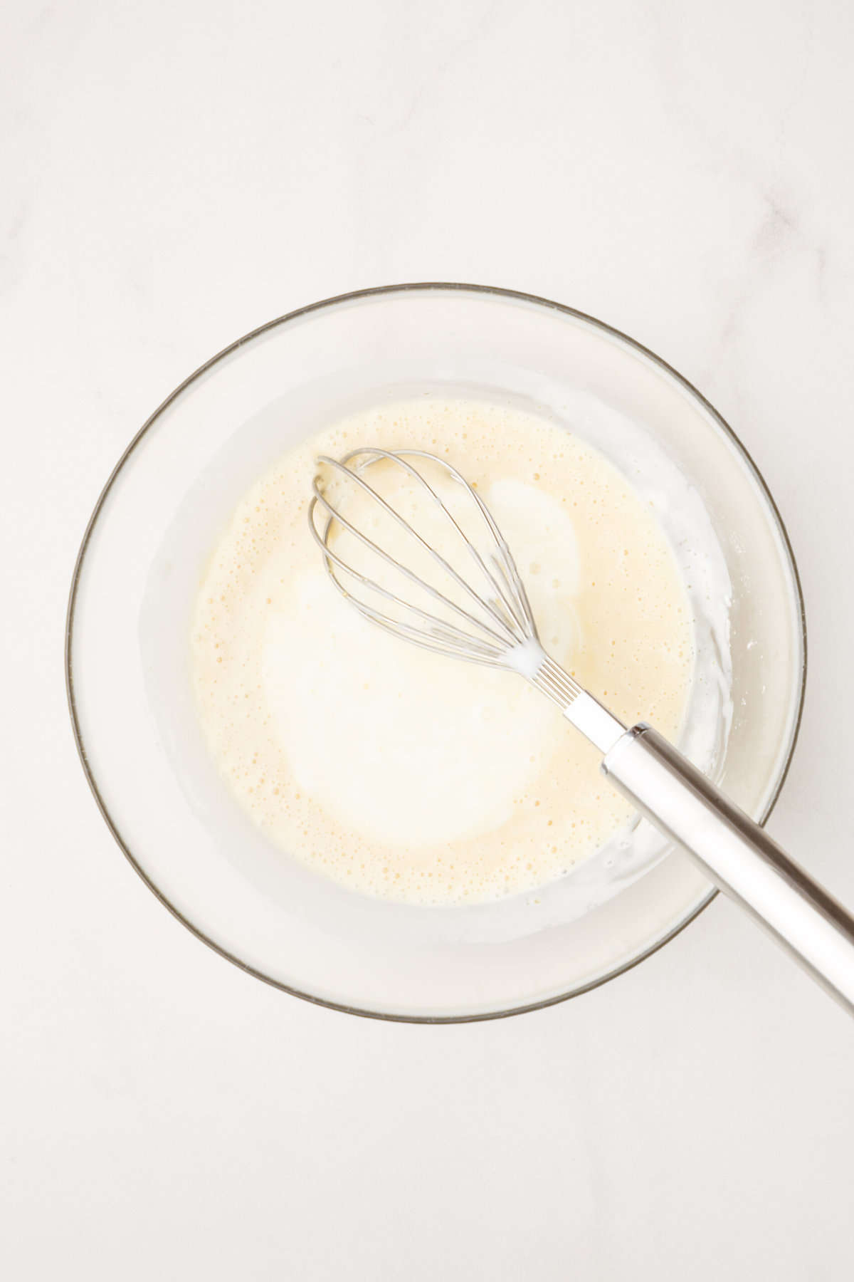 large glass bowl of cream, sugar and cornstarch with a metal whisk.