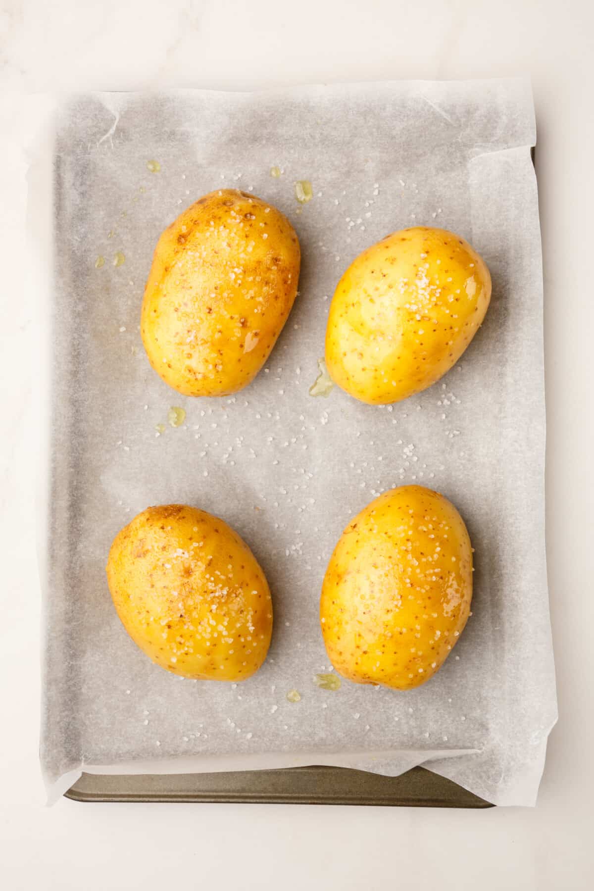 four russet potatoes pierced and salted sitting on a parchment-lined baking sheet.