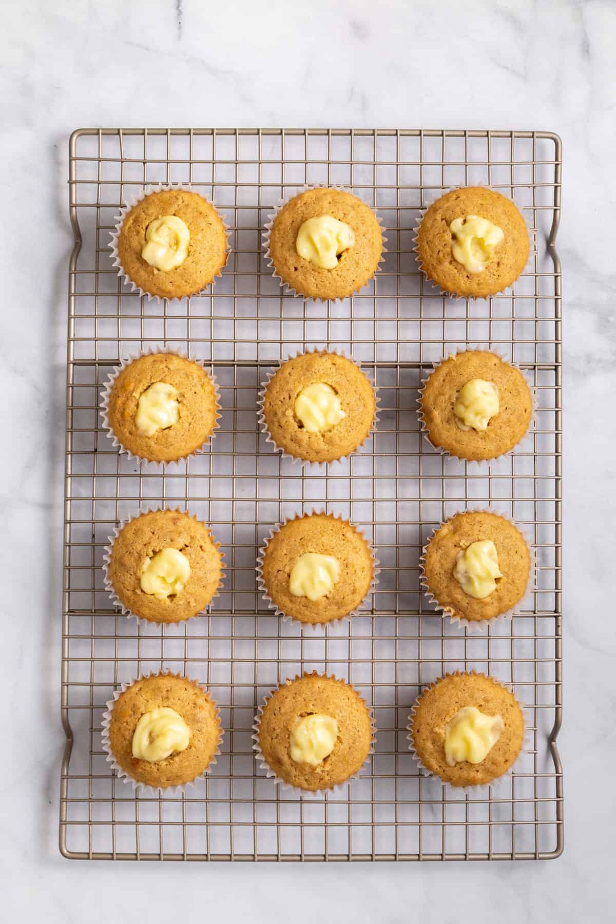 12 baked banana pudding cupcakes sitting on a cooling rack with banana pudding filling.
