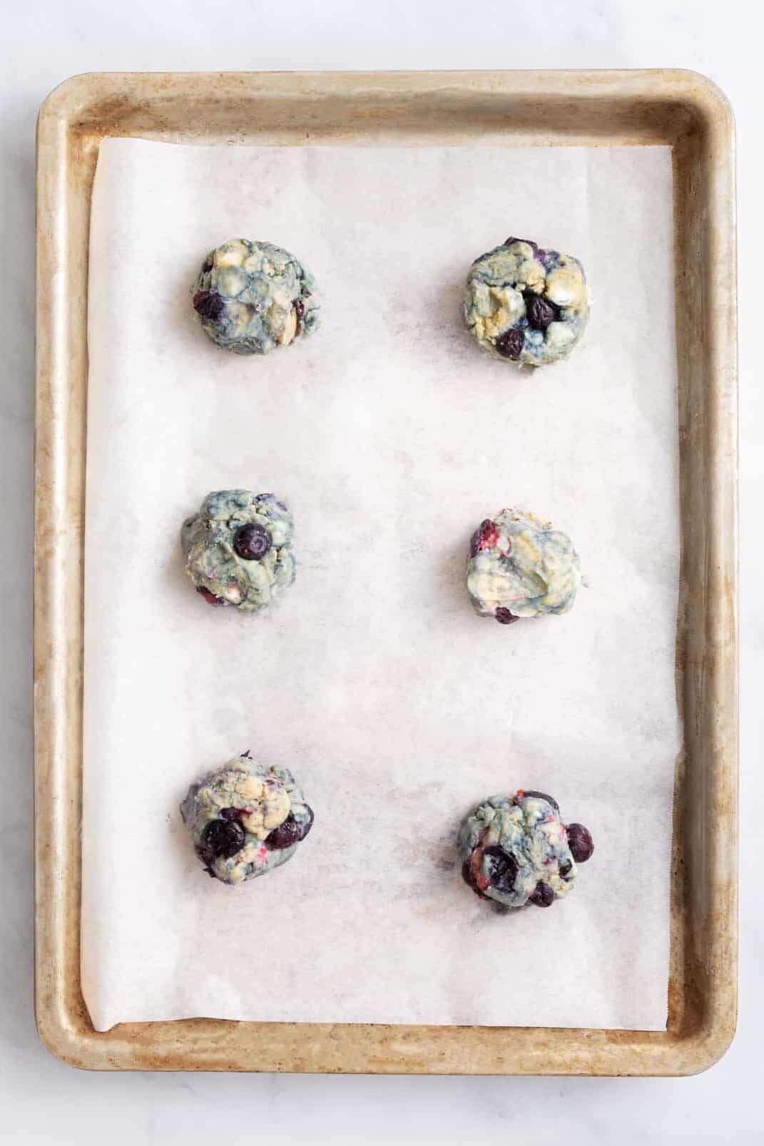 six arranged blueberry cookie dough balls on a parchment-lined baking sheet.