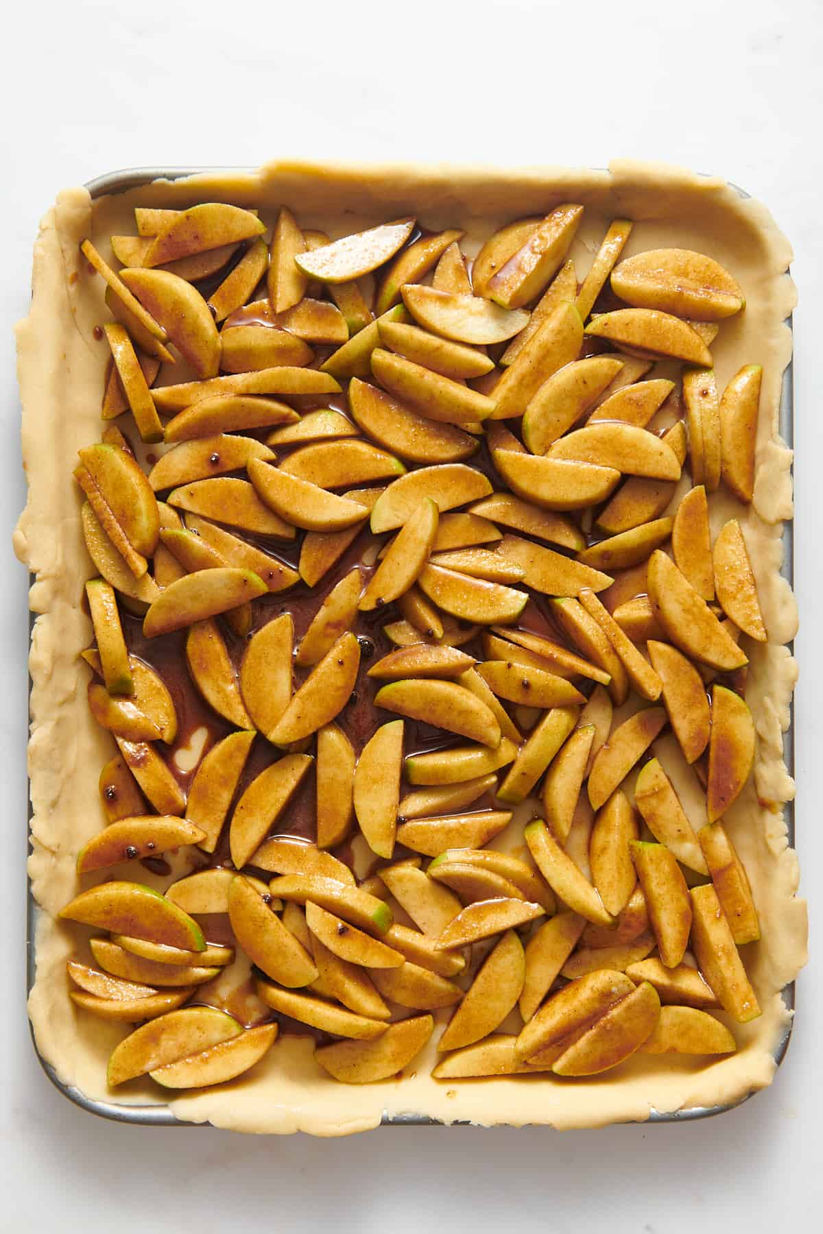 sliced green apples tossed in a cinnamon, brown sugar mixture spread evenly on a pie crust-lined baking tray.