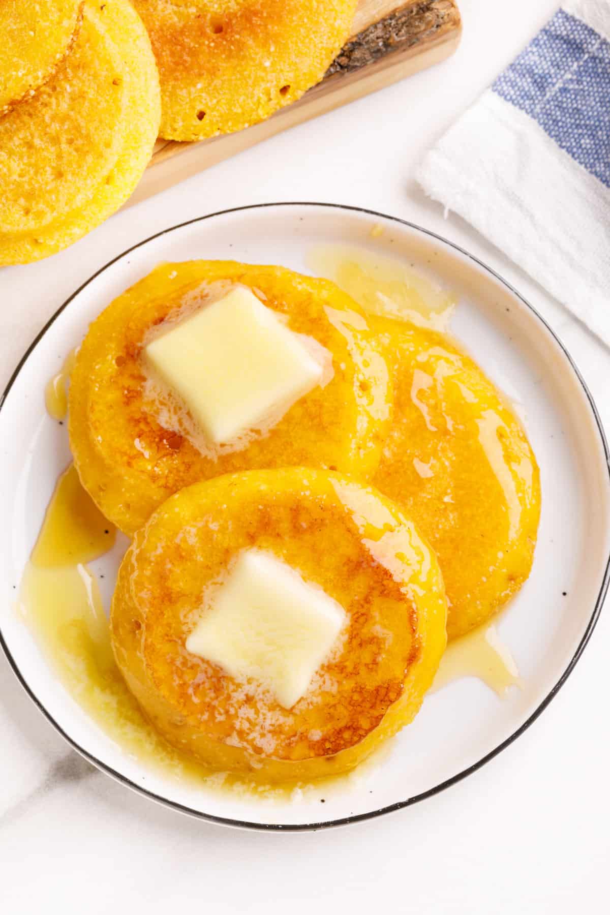 three cornbread fritters topped with butter and honey served on a white round plate. 
