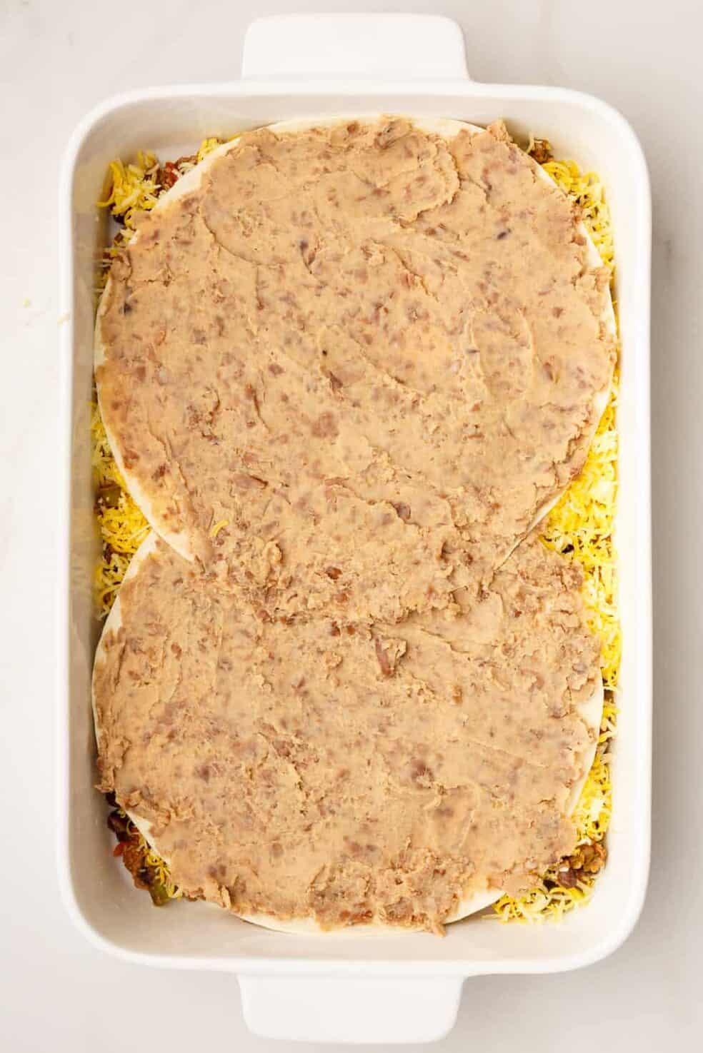 9x13 casserole dish with unbaked taco lasagna. the image shows two tortillas layered with smashed black beans.