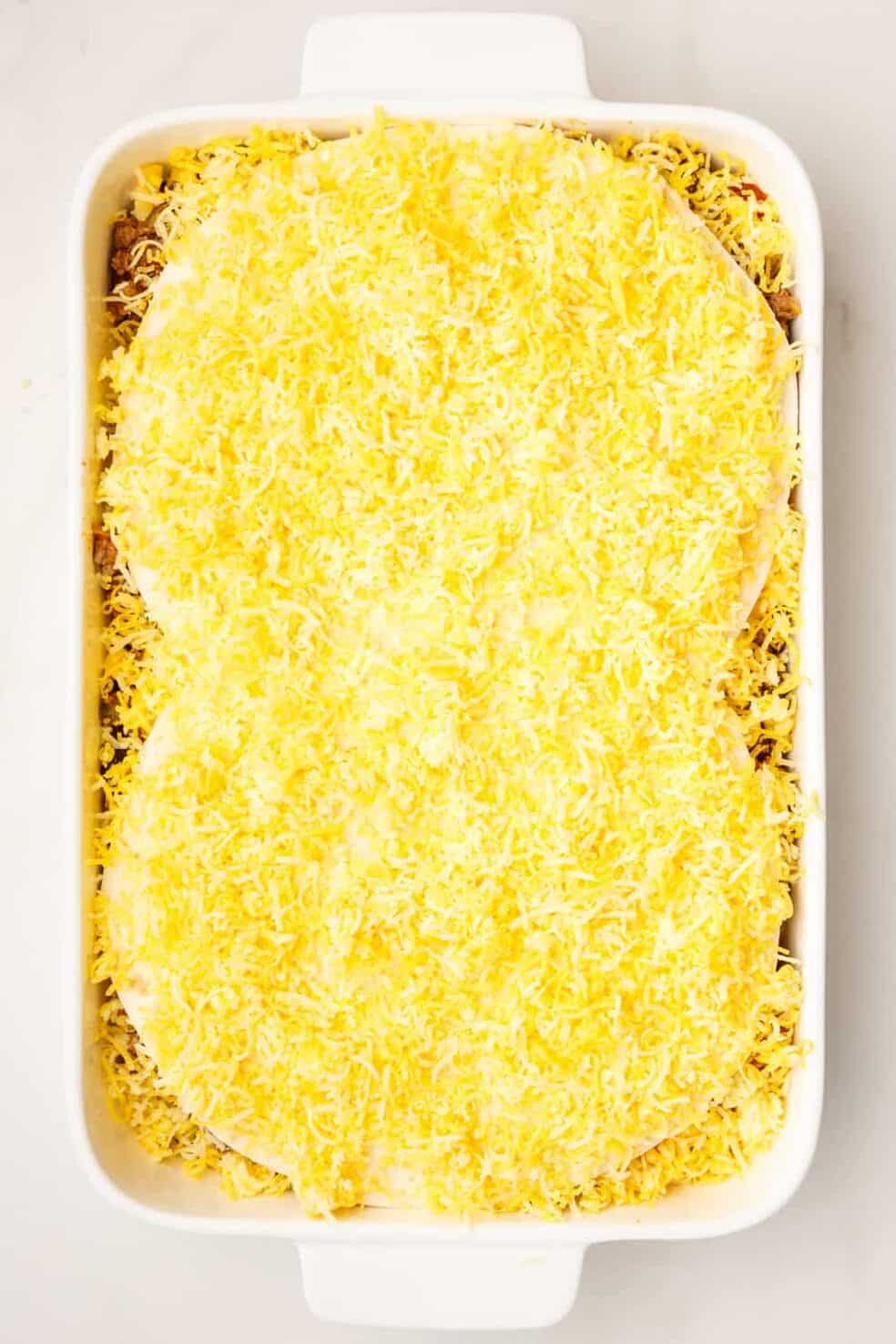 9x13 casserole dish with unbaked taco lasagna. the image shows two tortillas covered in shredded mexican cheese layered to the top.