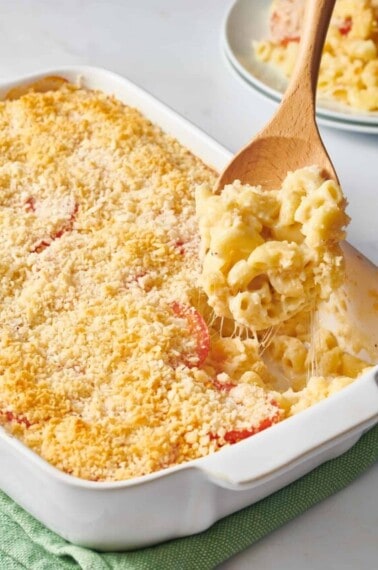 A wooden spoon lifting a scoop of Ina Garten mac and cheese from a baking dish.
