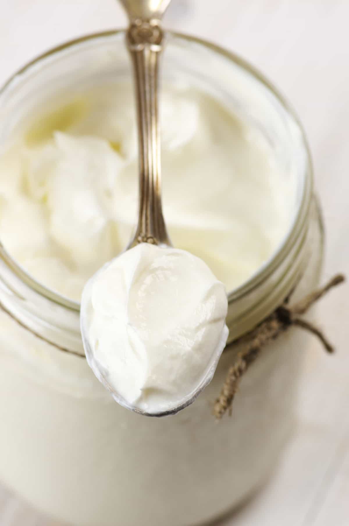 Homemade sour cream in glass jar with spoon close-up on rustic white wooden table.