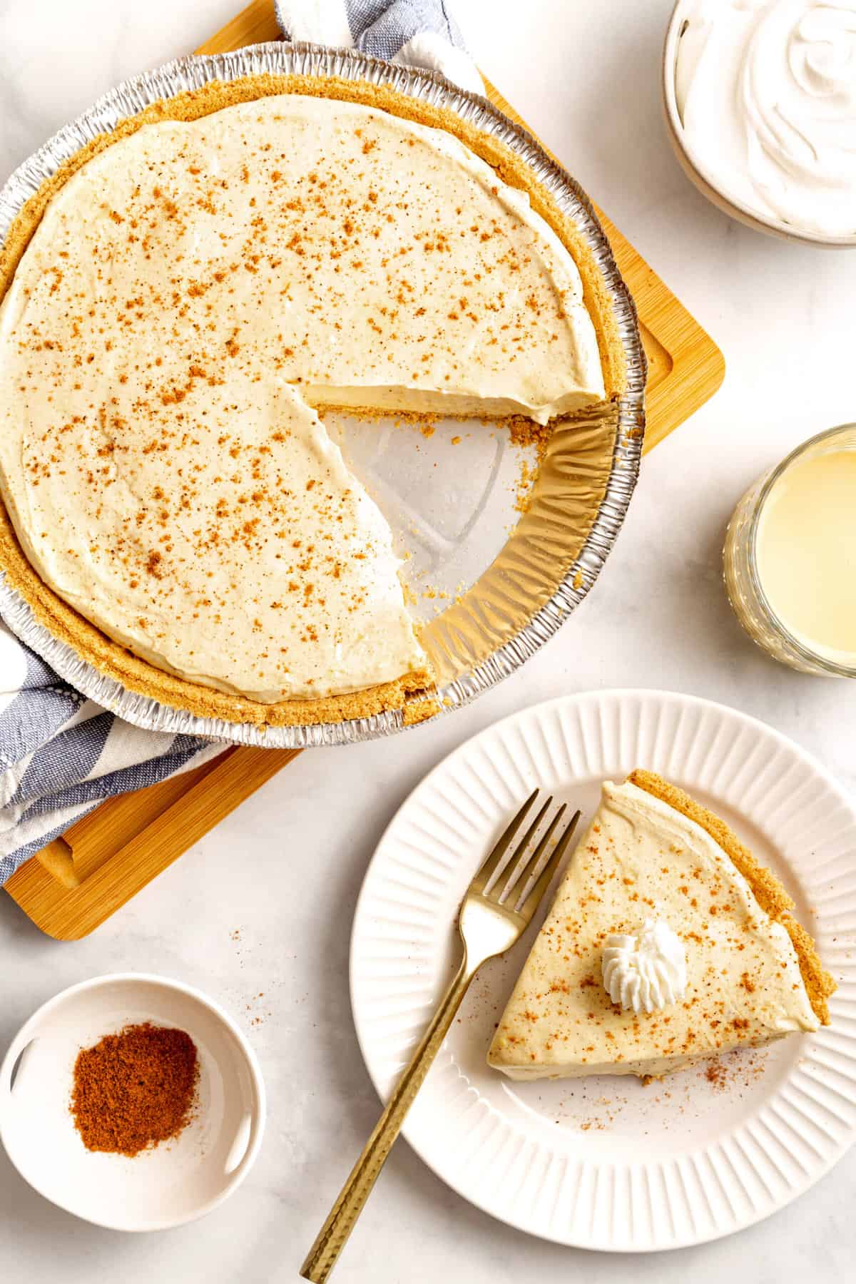 top down image of no bake egg nog pie and a slice served on a white round plate with a gold fork. 