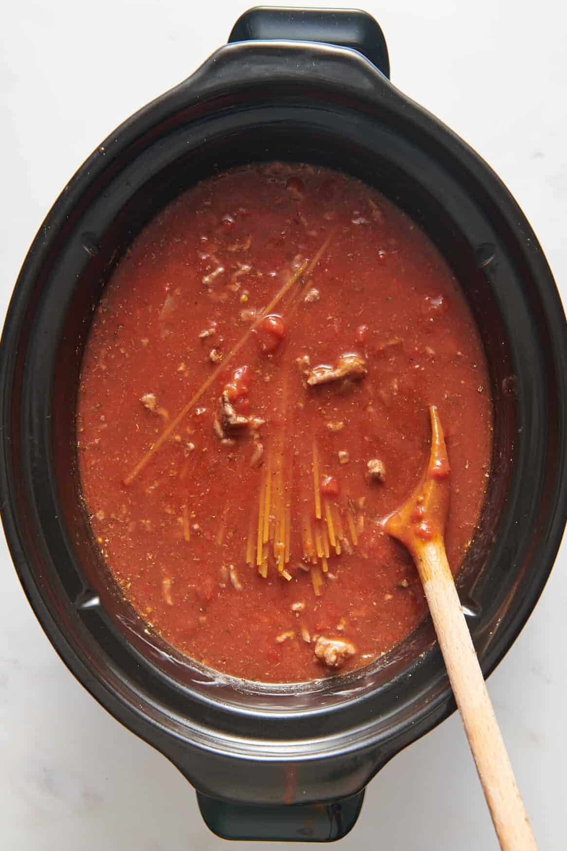 top down image of uncooked spaghetti added to the crock pot with tomato sauce and cooked ground beef. 