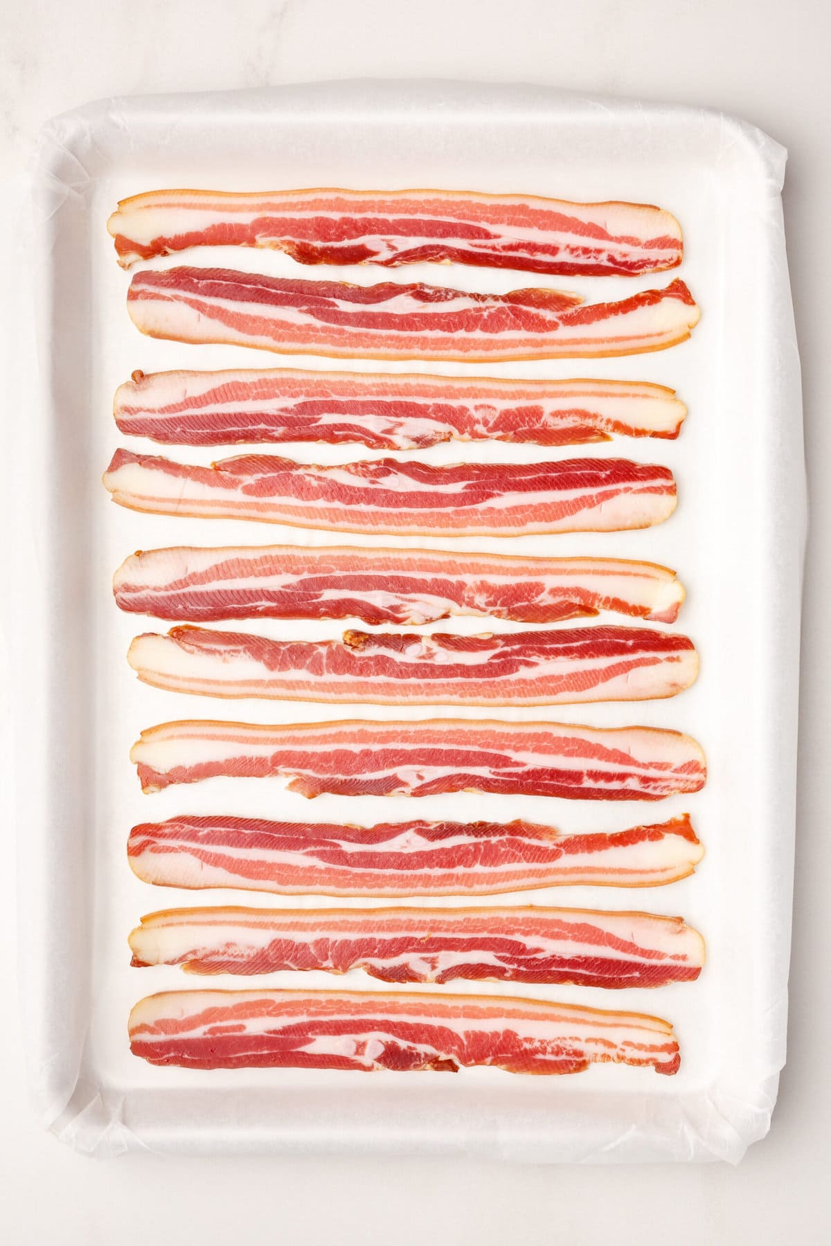 10 strips of uncooked bacon sitting on a parchment-lined baking sheet. 