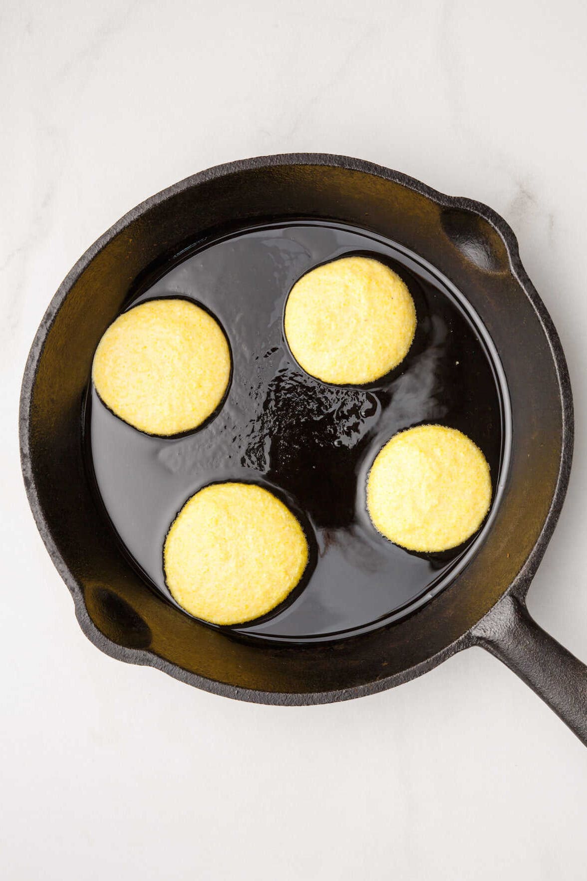 four cornbread fritters shallow frying on one side in a cast iron skillet. 